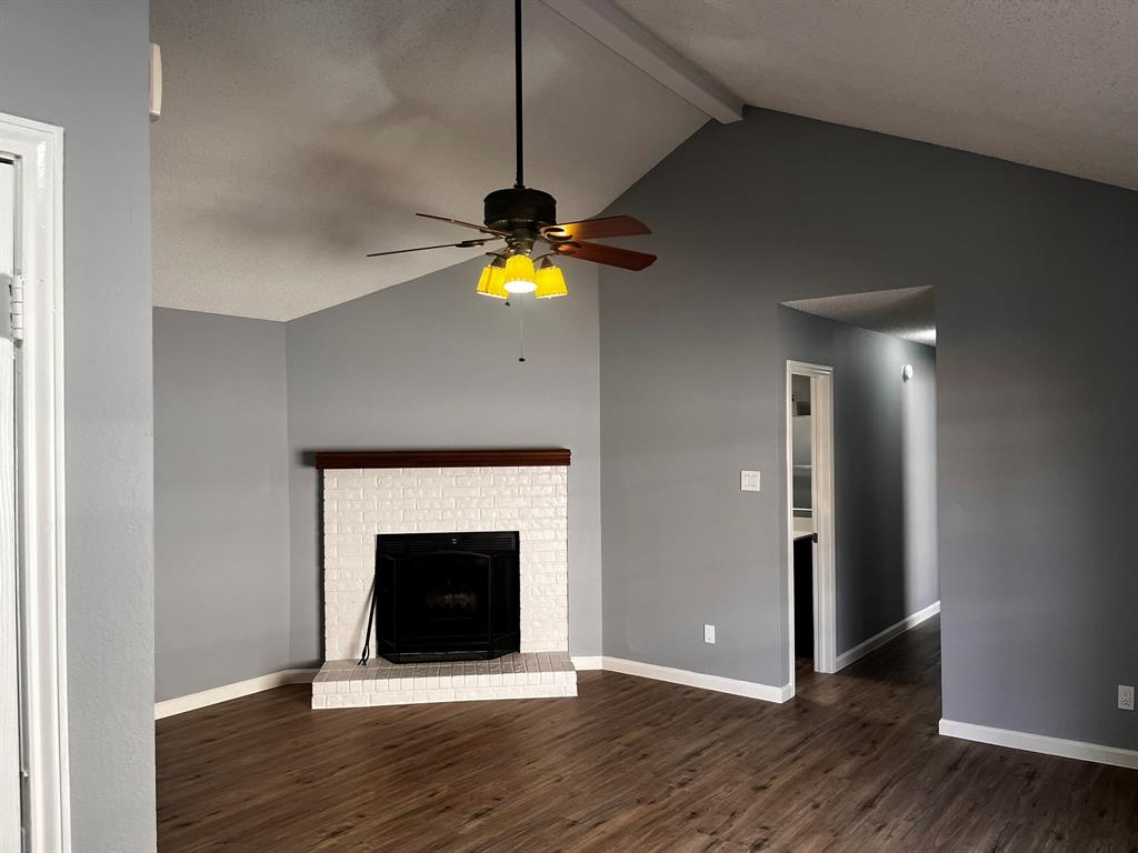 1007 Mack Place Denton, TX 76209 - Photo 12 of 37 Unfurnished living room with beamed ceiling, dark wood-style flooring, ceiling fan, a brick fireplace, and a textured ceiling
