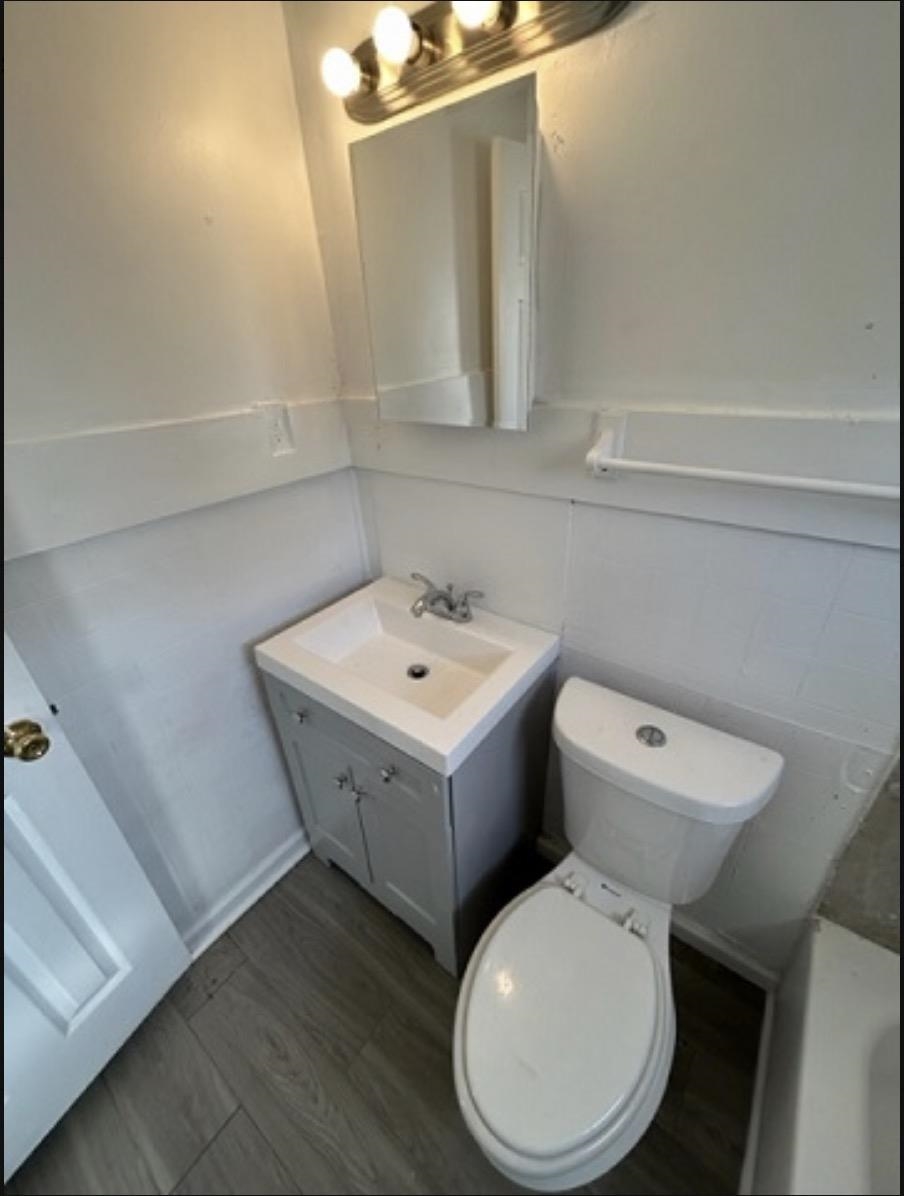 Compact bathroom featuring a white toilet, gray vanity with integrated sink, wood-finish flooring, and a mirrored medicine cabinet with overhead lighting