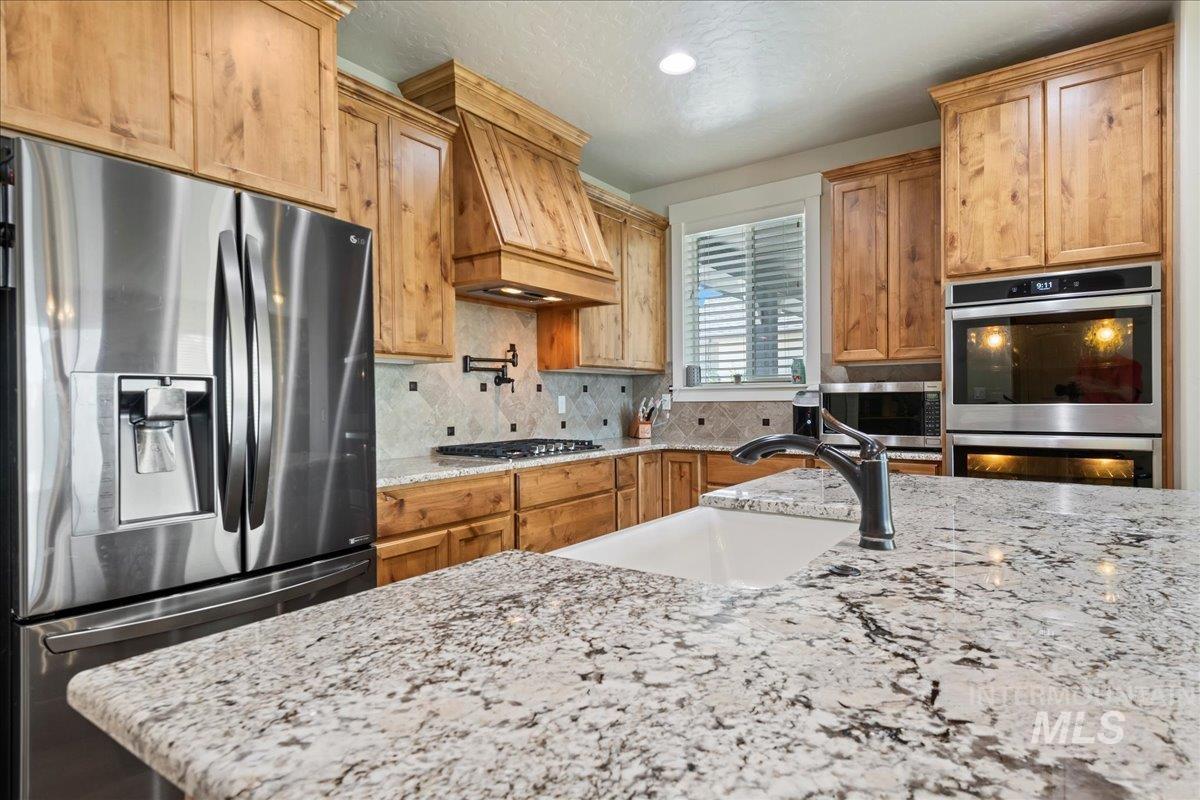 6091 East Path Drive Nampa, ID 83687 - Photo 12 of 33 Kitchen featuring appliances with stainless steel finishes, decorative backsplash, light stone counters, premium range hood, and brown cabinetry