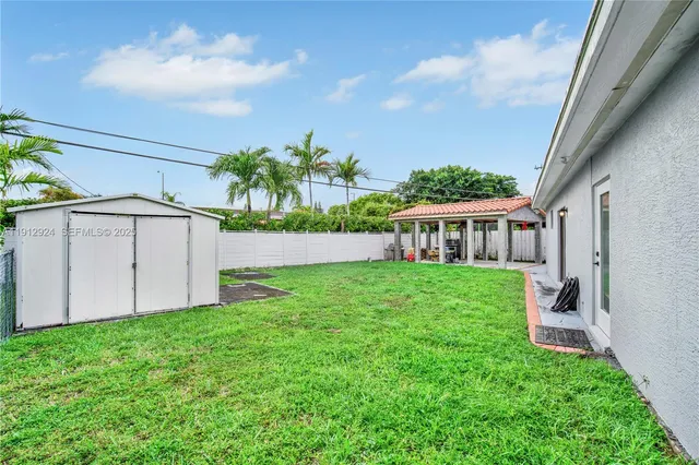 a view of a white house in front of a big yard with plants and large trees