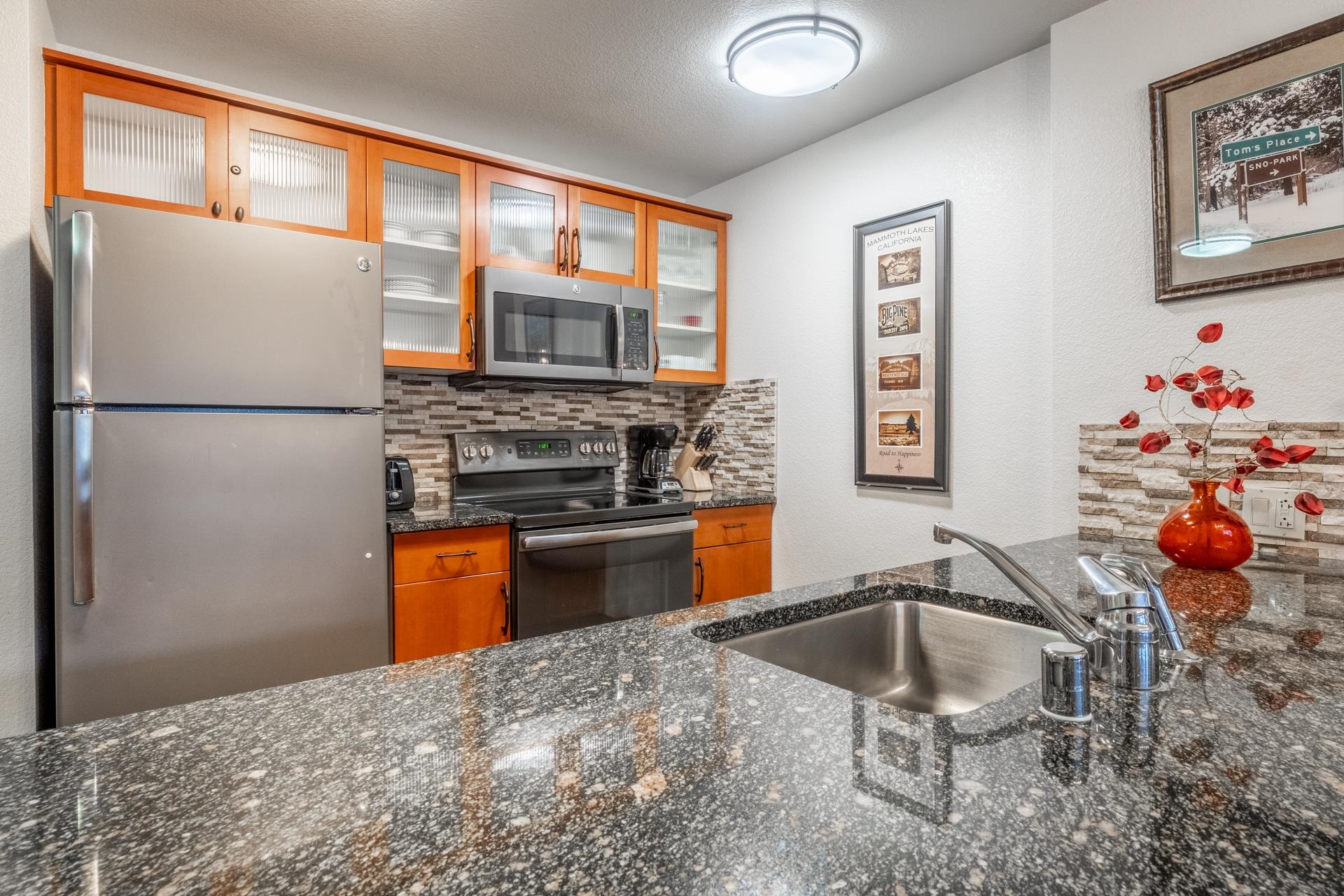6201 Minaret Road, Unit 2328 Mammoth Lakes, CA 93546 - Photo 4 of 23 Kitchen featuring stainless steel appliances, wood-finish cabinetry with glass inserts, a tile backsplash, and a black speckled countertop with an undermount sink