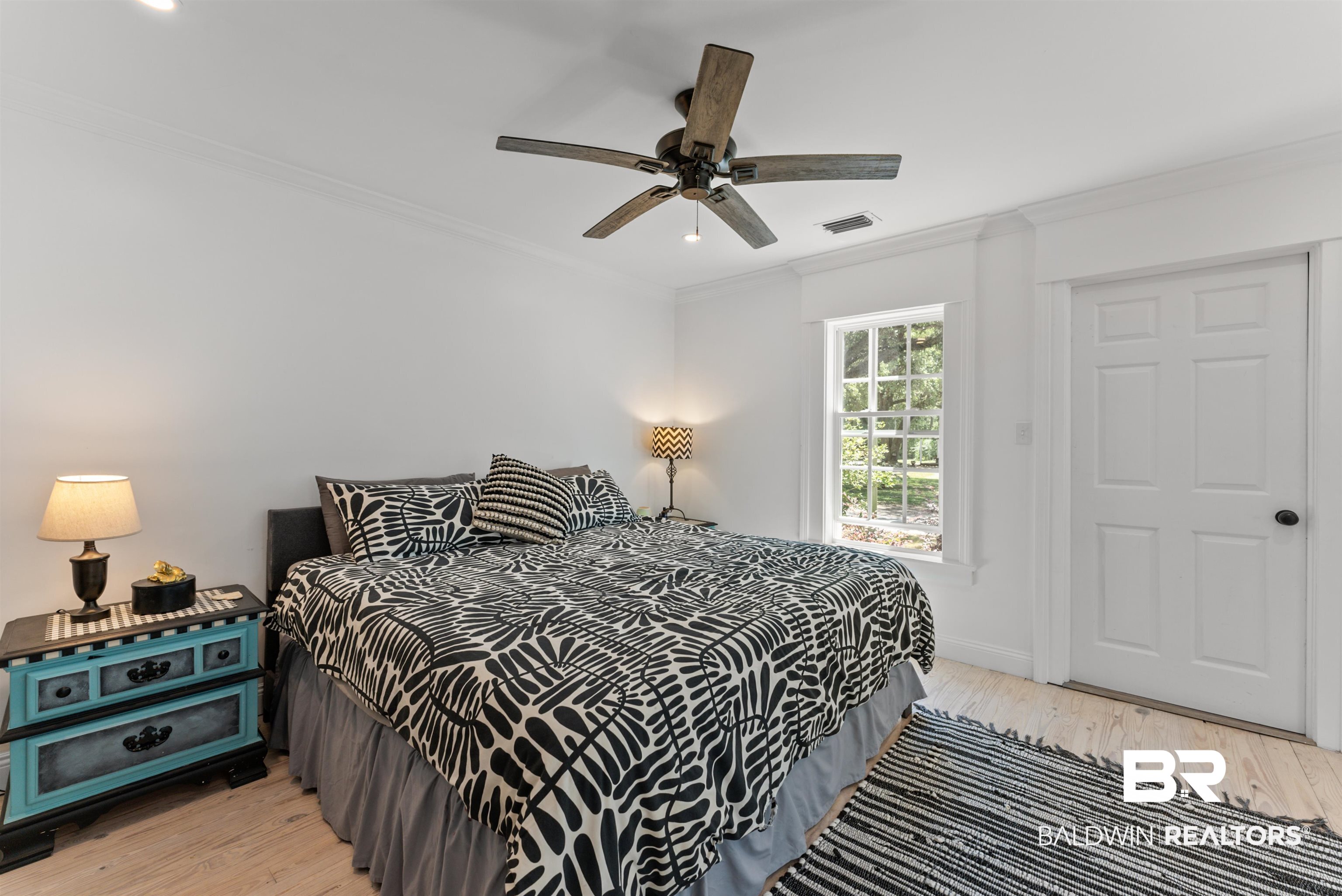 6447 Bemis Lane Bon Secour, AL 36511 - Photo 34 of 62 White walls and crown molding frame a bedroom featuring light wood-finish flooring, a ceiling fan, and a single window with a clear view of exterior greenery