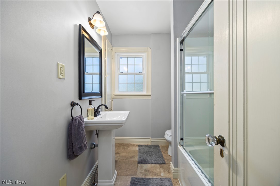 26019 Lake Road Bay Village, OH 44140 - Photo 19 of 40 Bathroom featuring a pedestal sink, framed mirror, wall-mounted lighting, a window with grid pattern, and a bathtub with a sliding glass enclosure