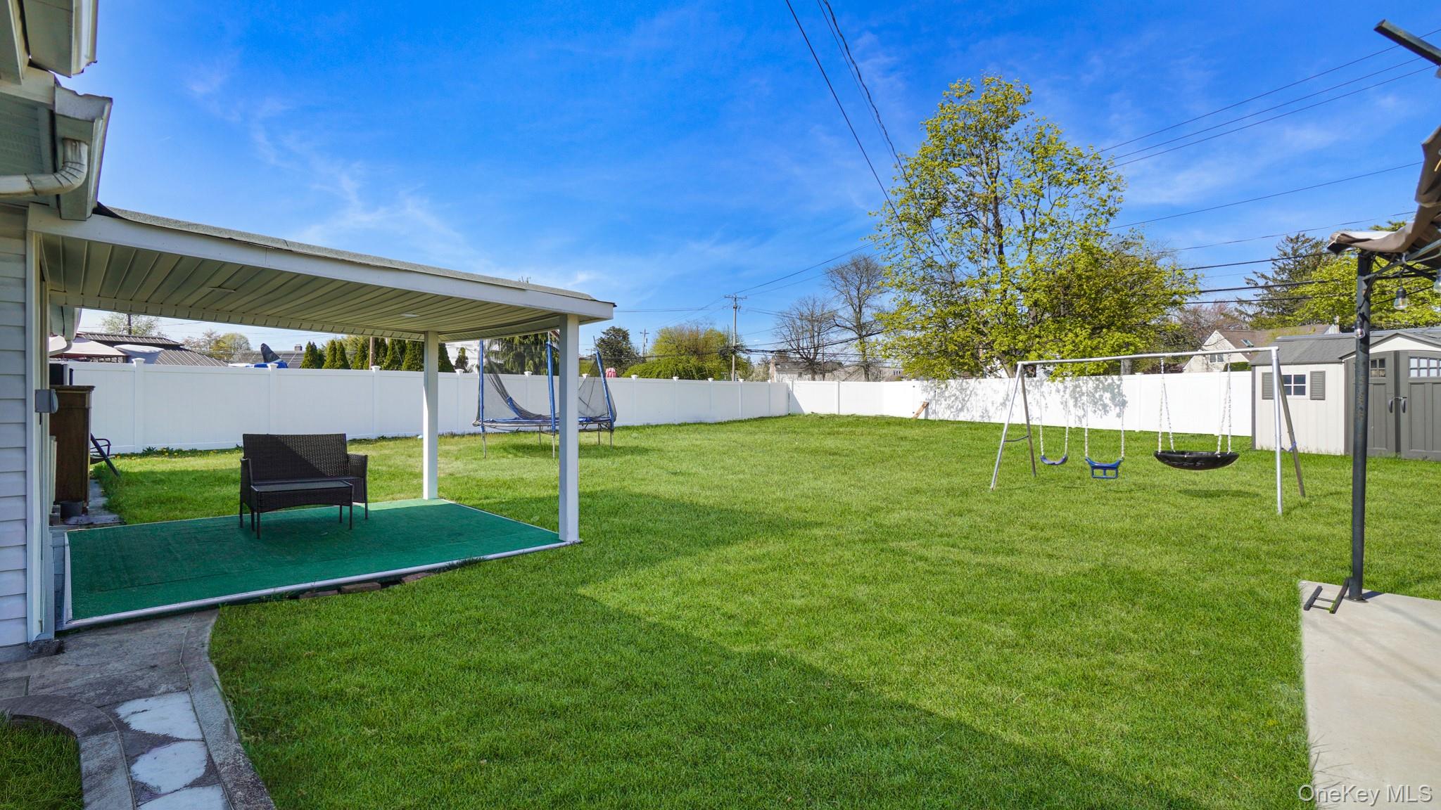 63 Blacksmith Road Levittown, NY 11756 - Photo 4 of 25 Expansive backyard featuring a covered patio with green outdoor carpeting, a large manicured lawn, a white privacy fence, and a storage shed