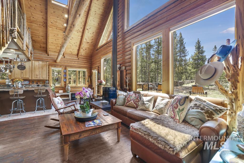 65 Goat Creek Way Stanley, ID 83278 - Photo 11 of 32 Vaulted great room featuring expansive windows with exterior tree views, exposed log beam construction, light wood-finish flooring, a wood-burning stove, and a kitchen pass-through with bar seating