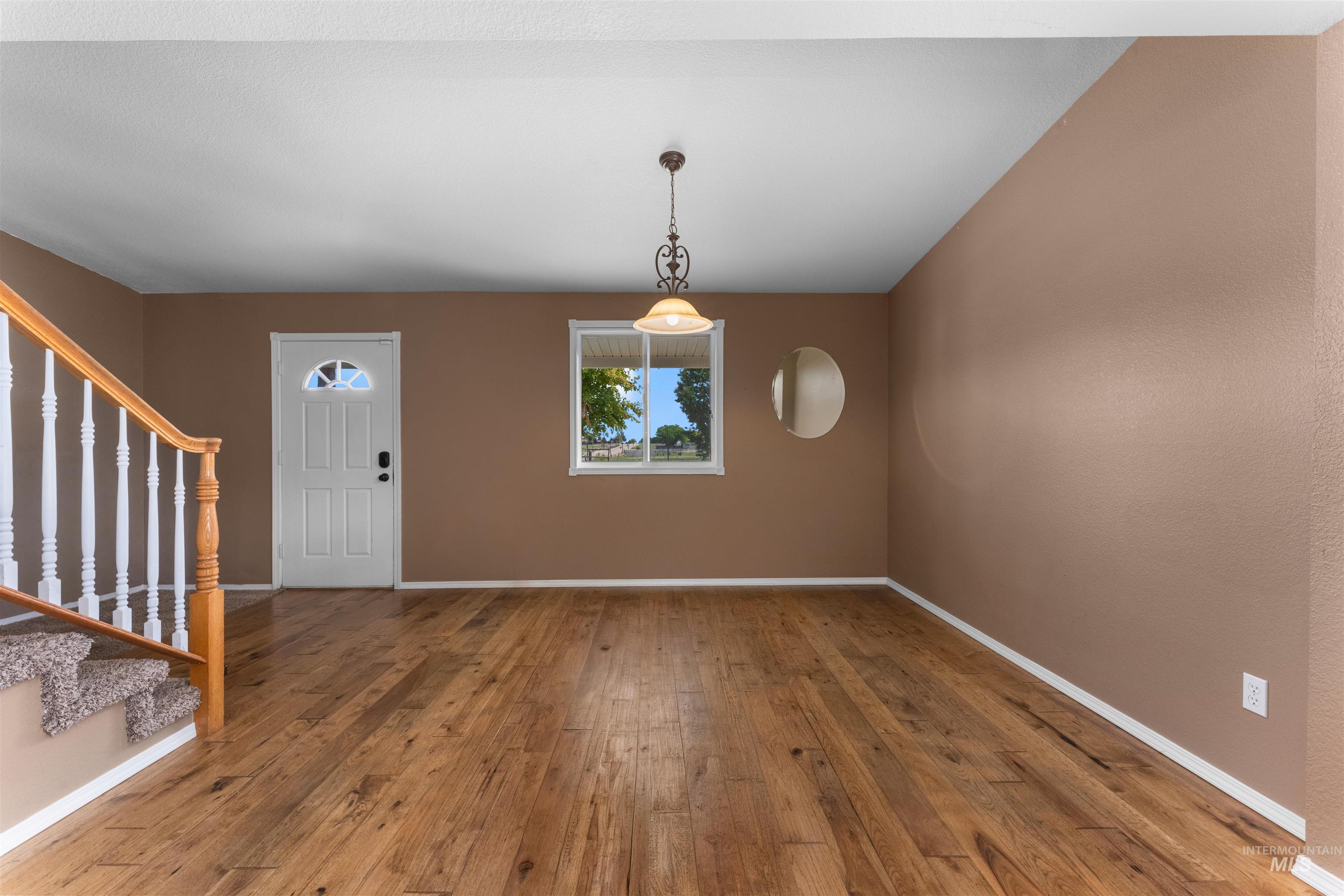 17441 Upper Pleasant Ridge Road Caldwell, ID 83607 - Photo 3 of 40 Spacious room featuring wood-finish flooring, a white paneled entry door with an arched window insert, a window providing natural light, a decorative ceiling-mounted light fixture, and a staircase with a wood handrail and carpeted steps