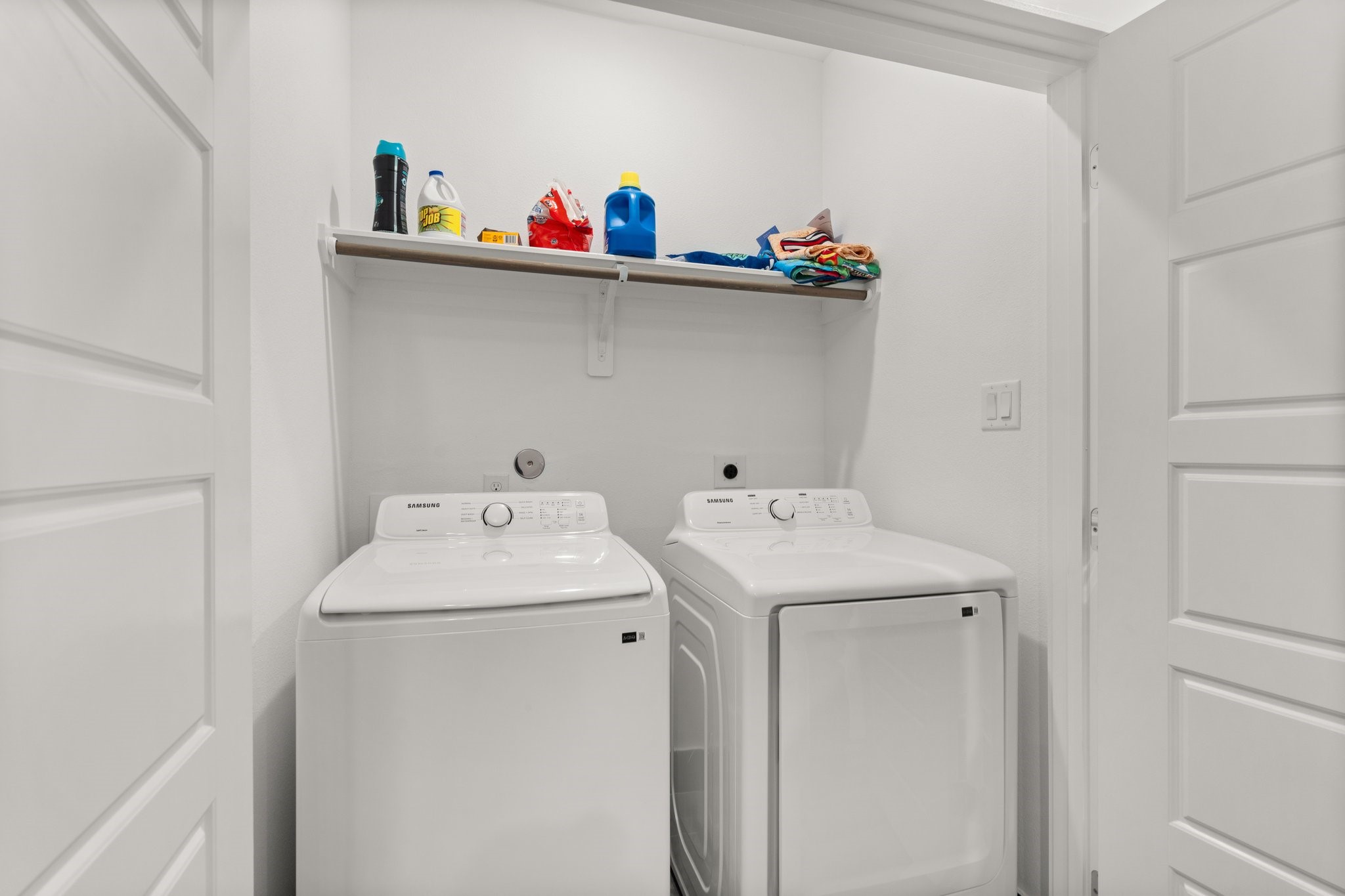 2928 Stevens Street Houston, TX 77026 - Photo 11 of 30 This photo shows a compact laundry area with white Samsung washer and dryer units. Above them is a shelf stocked with laundry supplies and towels, all set in a clean, white alcove with a door for easy access.