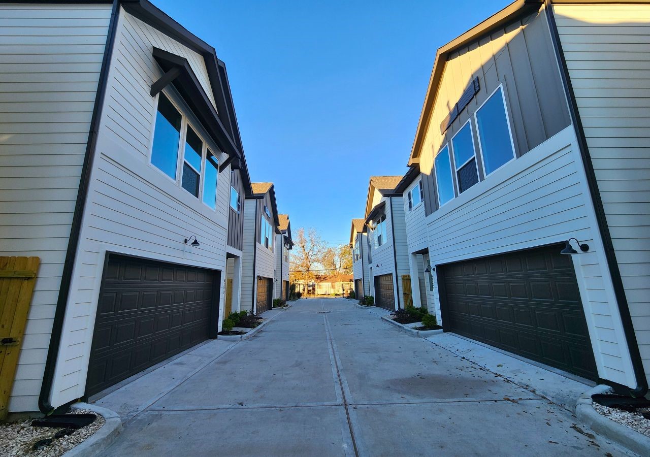 Modern townhouses with two stories, featuring clean white siding and dark garage doors. The homes are aligned along a paved driveway, creating a neat, organized community feel.