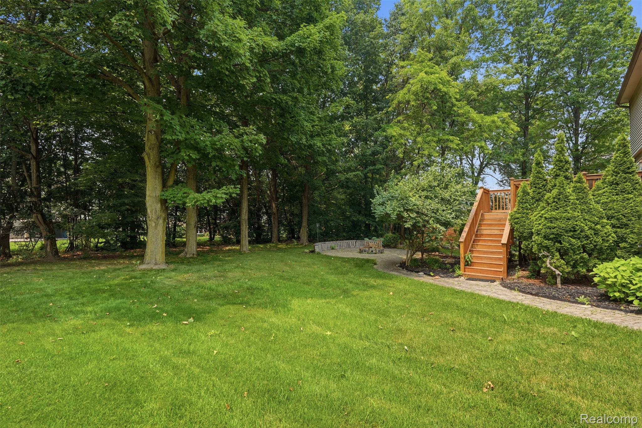 170 Fairhill Way Howell, MI 48843 - Photo 41 of 46 Expansive backyard featuring a lush lawn, mature trees, a wooden deck with stairs, a stone pathway, and landscaped garden beds