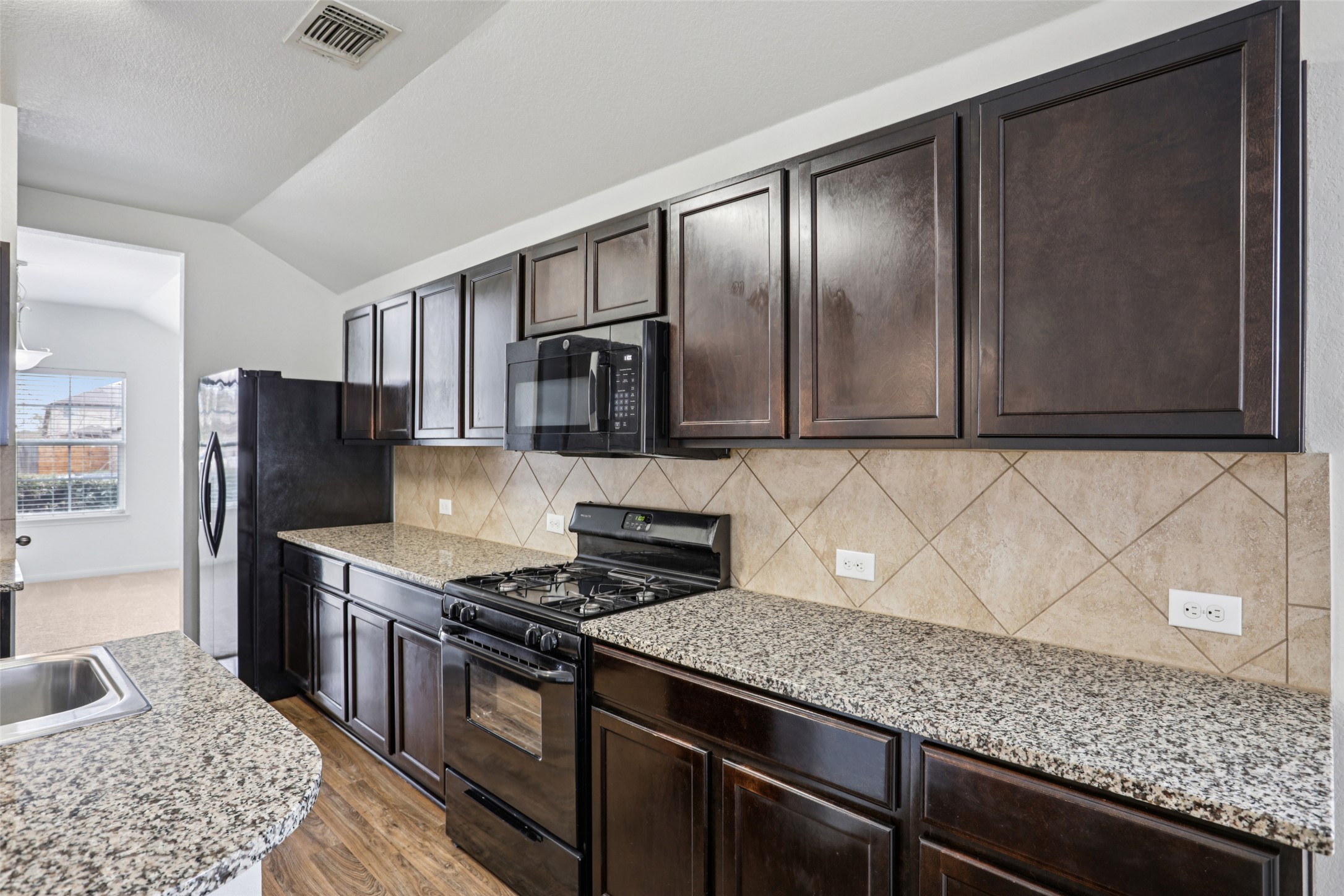 221 Alpha Kyle, TX 78640 - Photo 11 of 37 Kitchen with black appliances, dark wood finish cabinetry, decorative backsplash, vaulted ceiling, and light stone countertops