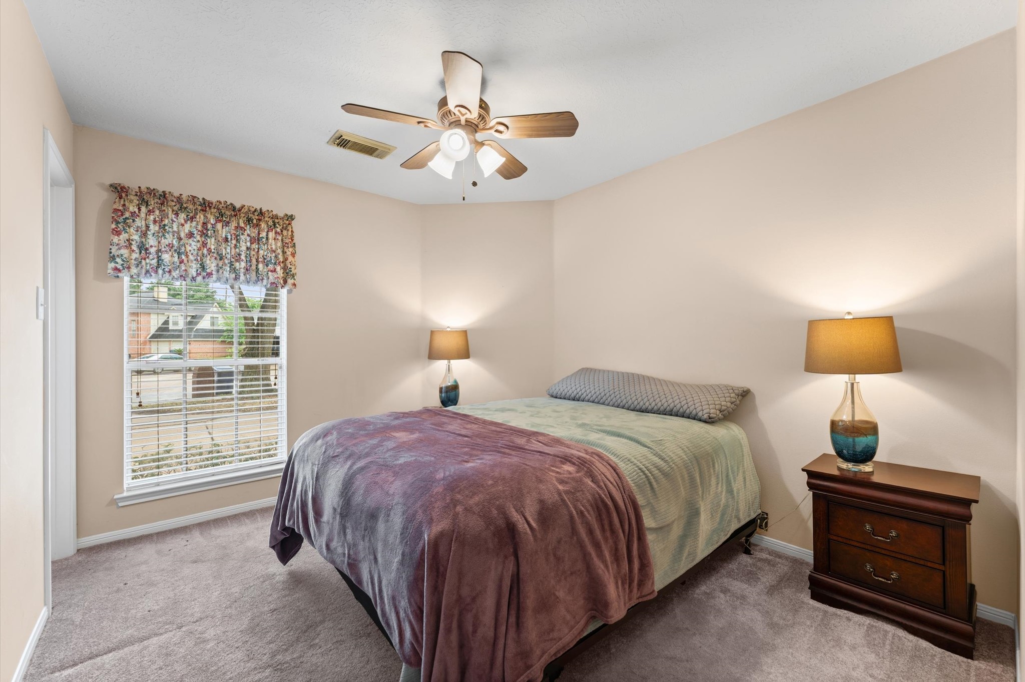 17618 Mill Springs Drive Spring, TX 77379 - Photo 16 of 26 This cozy bedroom features soft beige walls, a ceiling fan, and ample natural light from a large window with floral curtains. It's furnished with a comfortable bed, two matching bedside tables with lamps, and has plush carpeting for a warm, inviting feel.