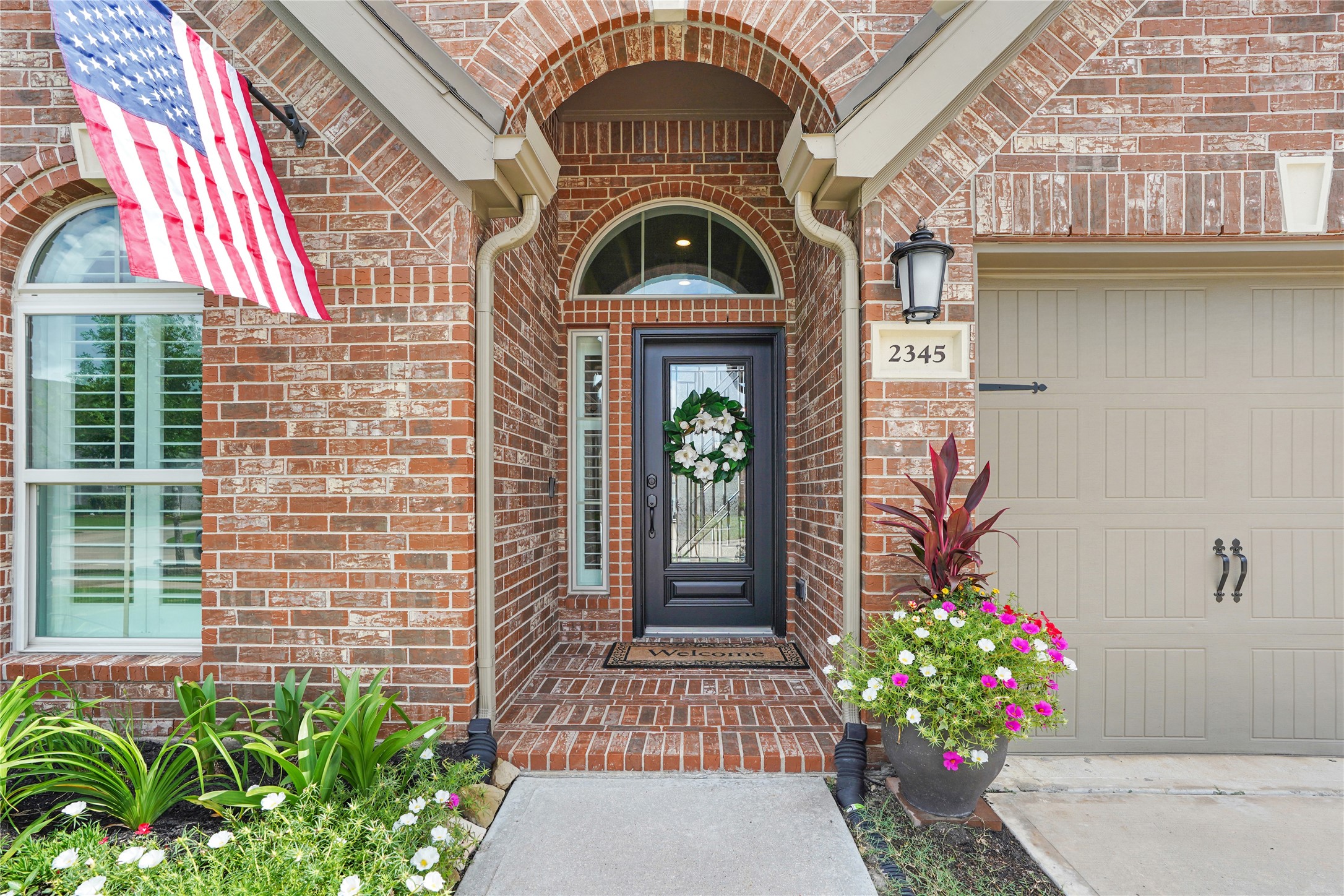 2345 Redwood Ridge Trail Manvel, TX 77578 - Photo 3 of 39 This charming entry features a welcoming brick facade with an arched doorway, beautifully accented by a black front door and decorative wreath. The entry is flanked by vibrant flower pots and neatly landscaped greenery, offering a warm and inviting first impression. An American flag adds a touch of classic charm.