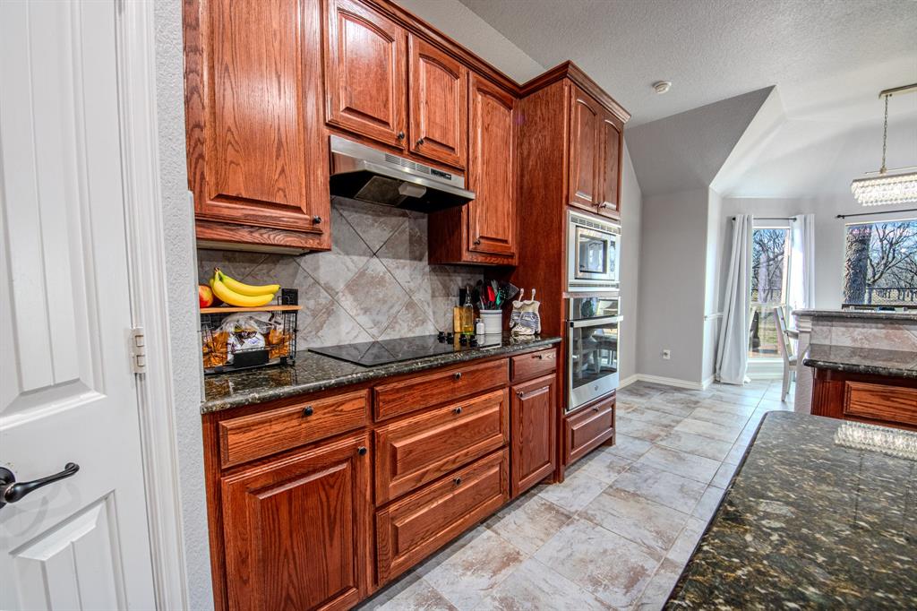 9001 Ravenswood Road Granbury, TX 76049 - Photo 15 of 38 Kitchen with dark stone countertops, stainless steel appliances, wood finish cabinets, pendant lighting, and backsplash