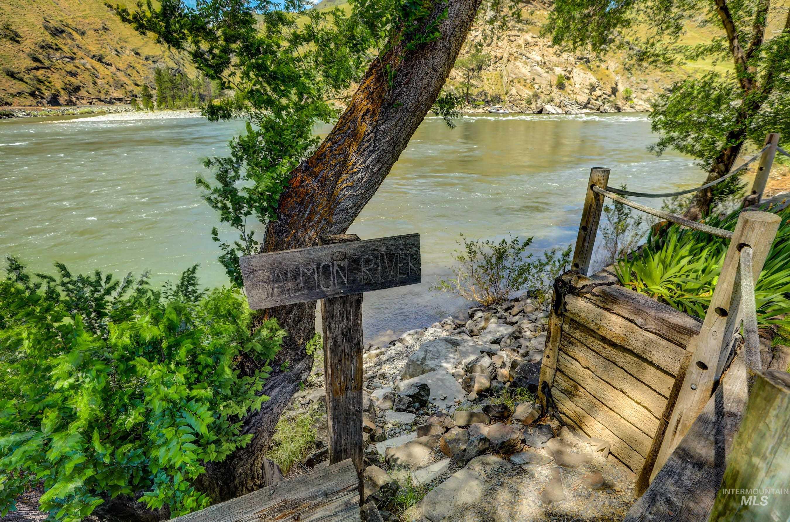 1432 Salmon Drive Riggins, ID 83549 - Photo 38 of 39 Riverside property featuring a wooden sign, natural wood railings, mature trees, and lush green foliage bordering a flowing river