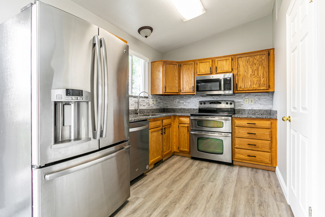4239 West 5325th Street South Roy, UT 84067 - Photo 7 of 26 Kitchen featuring wood-finish cabinetry, stainless steel appliances, granite-style countertops, mosaic tile backsplash, and light wood-finish flooring
