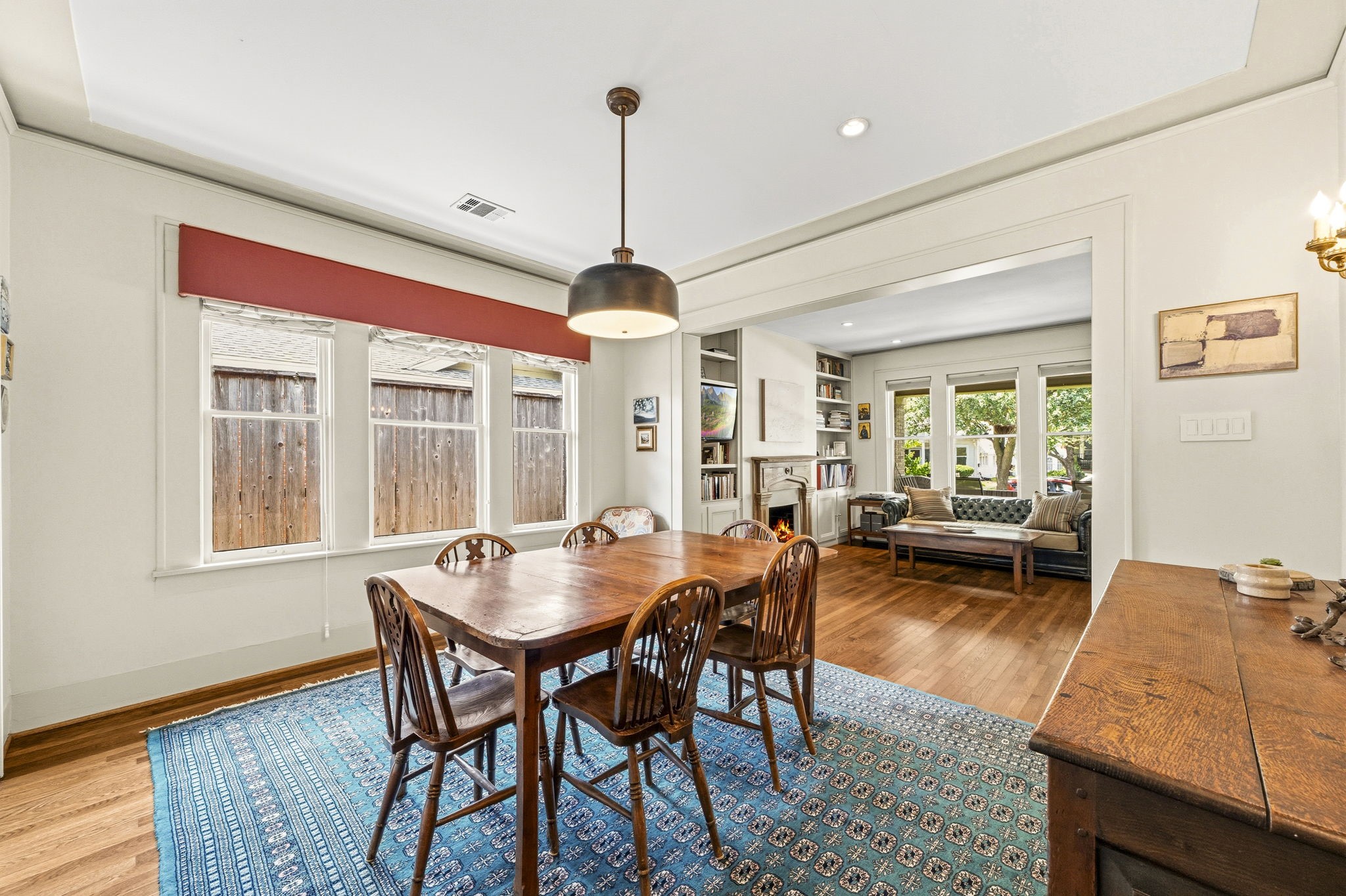 1521 Kipling Street Houston, TX 77006 - Photo 12 of 35 Bright dining area with wooden floors, a cozy rug, and a classic chandelier. Adjacent living space features a fireplace, built-in shelves, and large windows offering natural light and garden views.