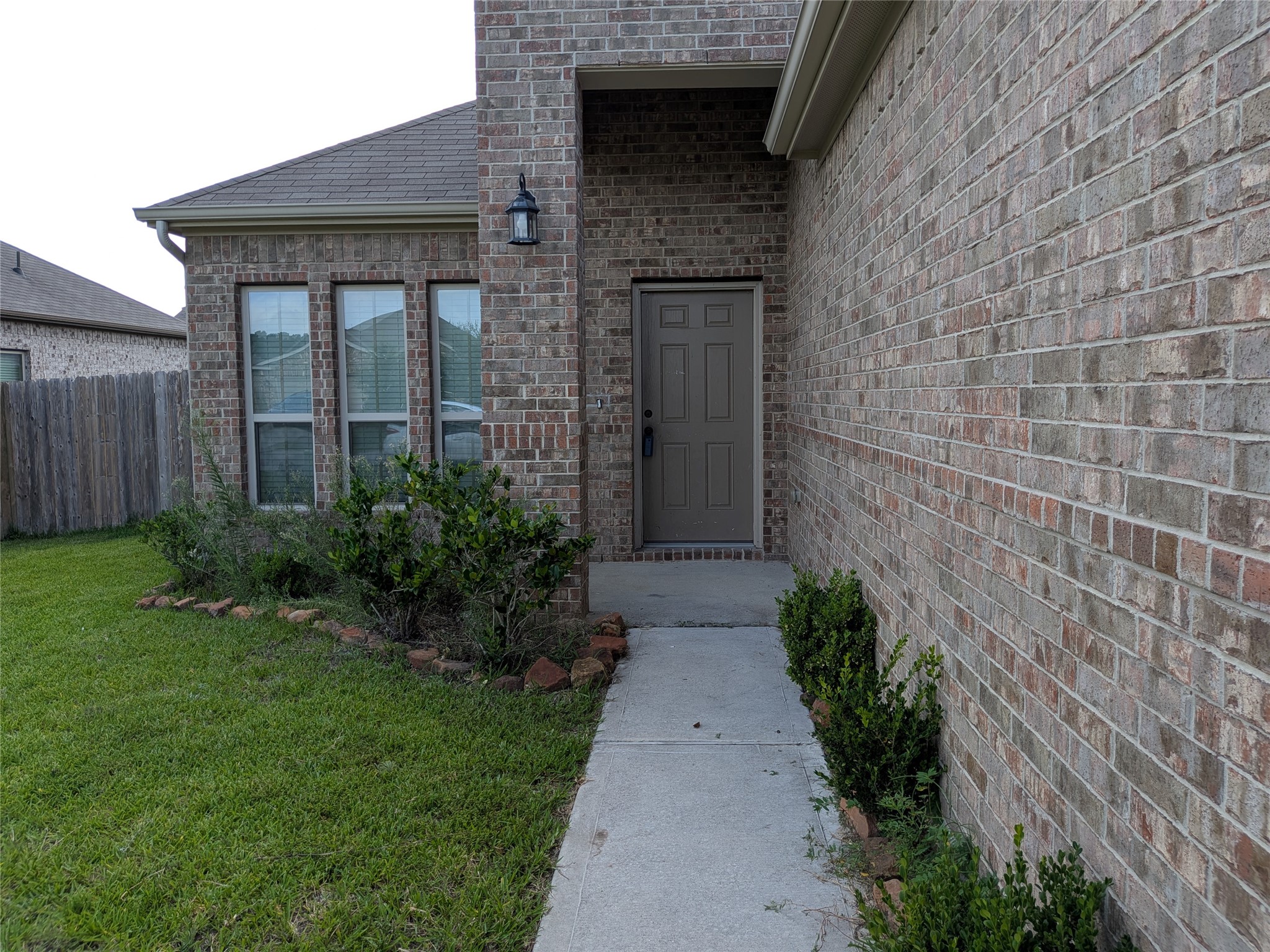 15730 Joe Dimaggio Street Splendora, TX 77372 - Photo 1 of 20 This photo shows the front entryway of a brick home, featuring a modest porch with a single door and outdoor light. A concrete path is flanked by tidy shrubbery, and a wooden fence borders the lawn on the left.