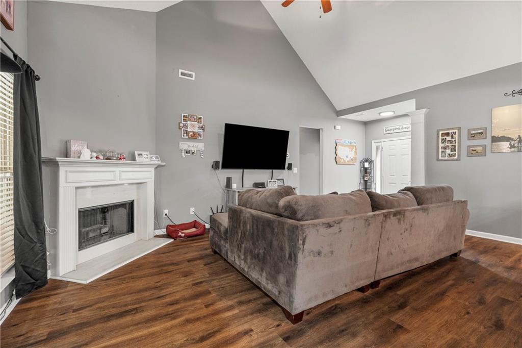 Living area featuring high vaulted ceiling, dark wood finished floors, a ceiling fan, a fireplace with raised hearth, and baseboards
