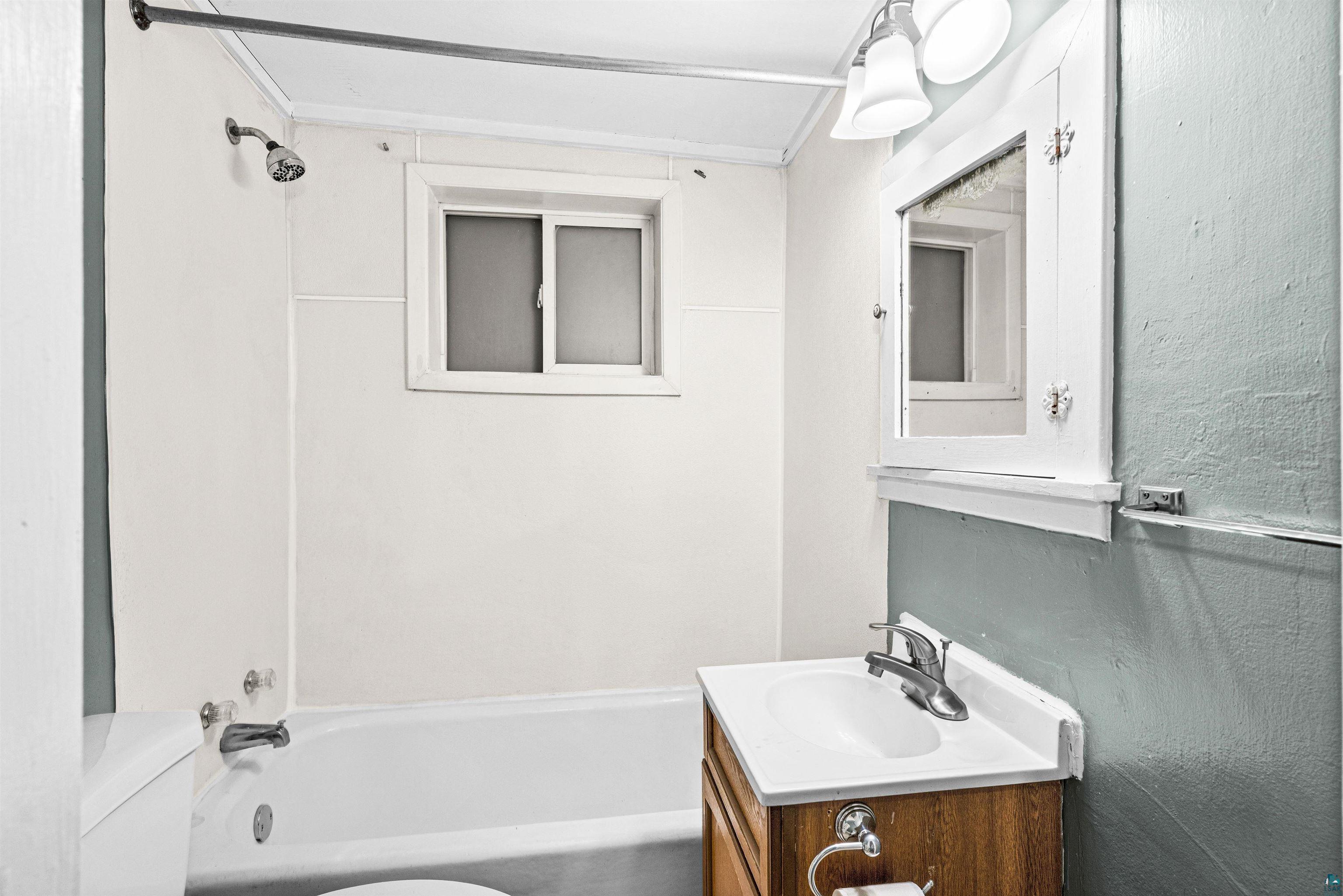 1127 East 3rd Street Duluth, MN 55805 - Photo 21 of 28 Bathroom featuring a white bathtub, shower fixture, sliding window, wood-finish vanity with an integrated sink, and a wall-mounted medicine cabinet with integrated lighting