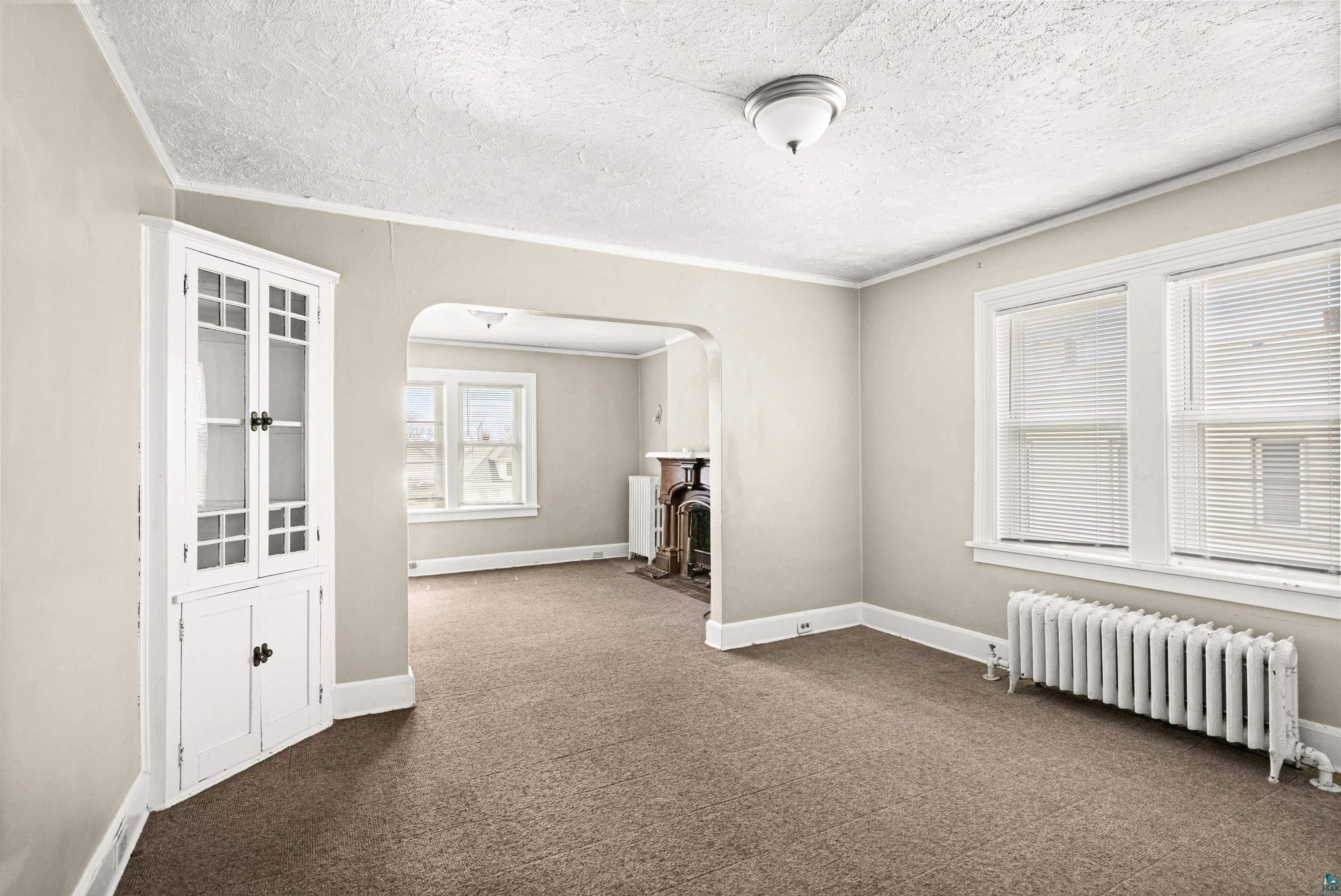 1127 East 3rd Street Duluth, MN 55805 - Photo 22 of 28 Neutral-toned room with carpet flooring featuring a built-in corner cabinet with glass-front doors, multiple windows, a ceiling-mounted light fixture, and a radiator