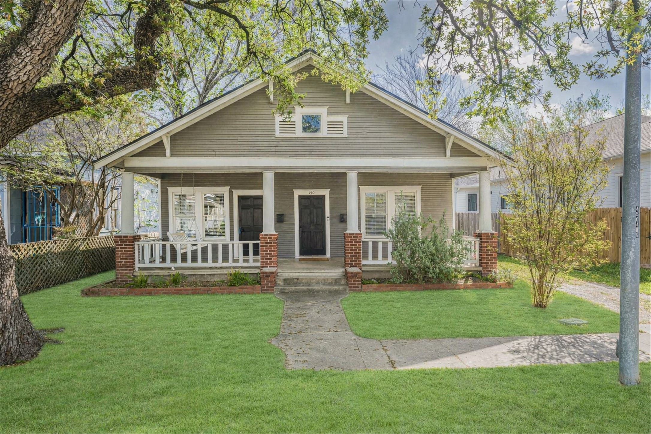 Charming bungalow-style home with a welcoming front porch and swing, surrounded by lush greenery. The exterior features classic brick accents, a tidy lawn, and a cozy, inviting entrance. Perfect for enjoying quiet neighborhood living.