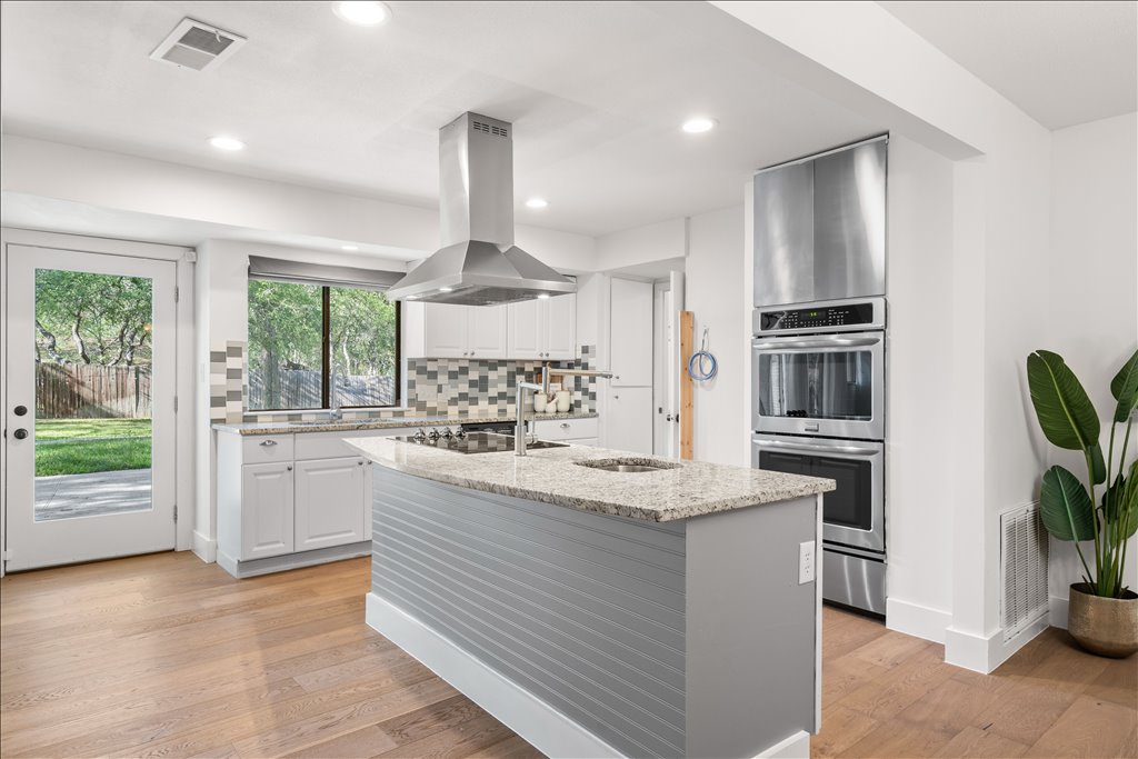 3402 Saddlestring Trail Austin, TX 78739 - Photo 17 of 33 Kitchen featuring light stone countertops, decorative backsplash, island exhaust hood, stainless steel double oven, and light wood-style flooring