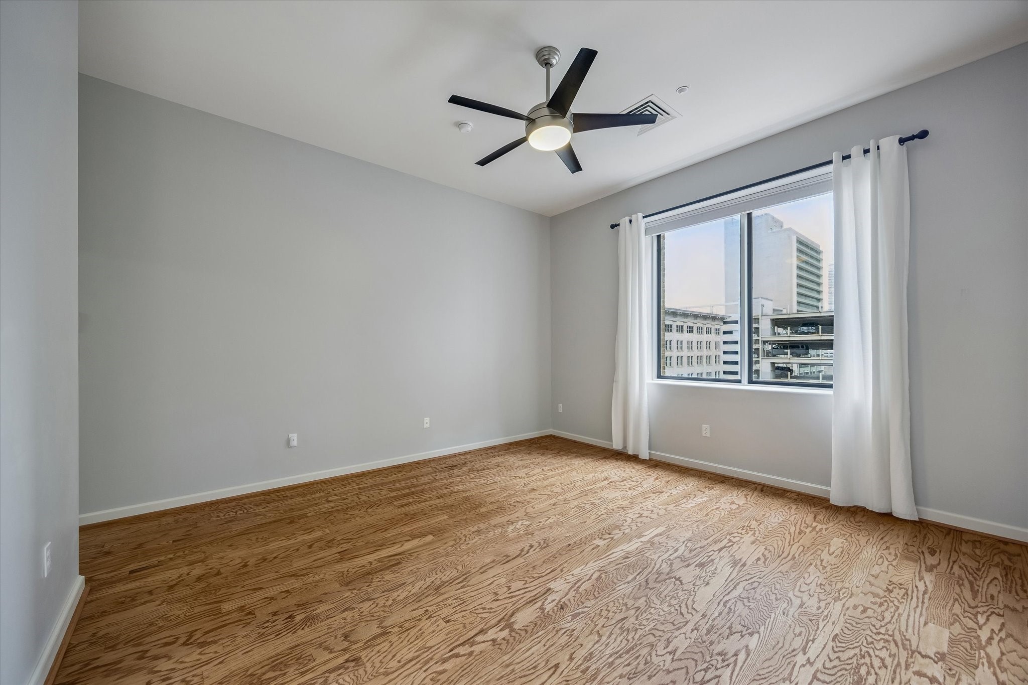 914 Main Street, Unit 910 Houston, TX 77002 - Photo 10 of 32 Primary bedroom features hardwood floors, a large window with city views, white curtains, and a modern ceiling fan, offering a bright and airy atmosphere.