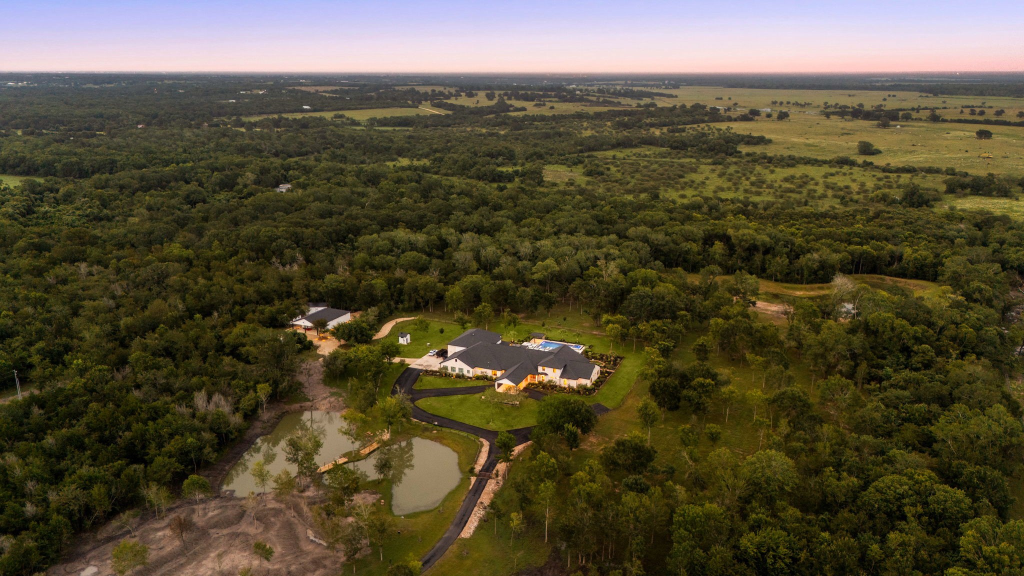 1100 Felder Road Washington, TX 77880 - Photo 45 of 50 This sweeping aerial view captures the estate's setting at dusk, with the main residence, additional outbuilding, and two ponds nestled among mature trees — offering a rare blend of privacy.