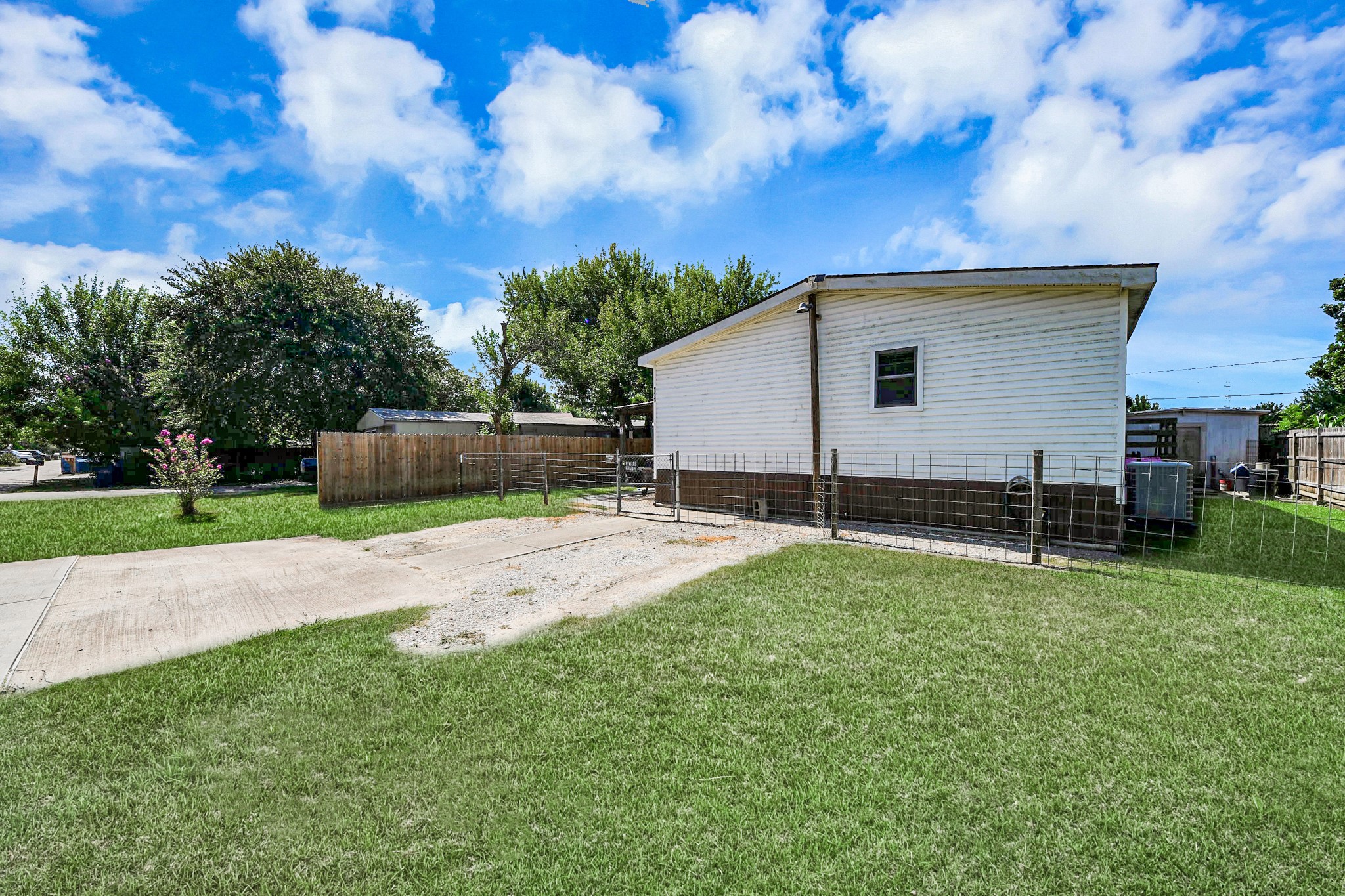 131 Kiowa Court Rosharon, TX 77583 - Photo 30 of 42 A single-story manufactured house with white siding, set on a spacious grassy lot. There's a paved driveway and a fenced-in area, surrounded by trees and a clear blue sky.