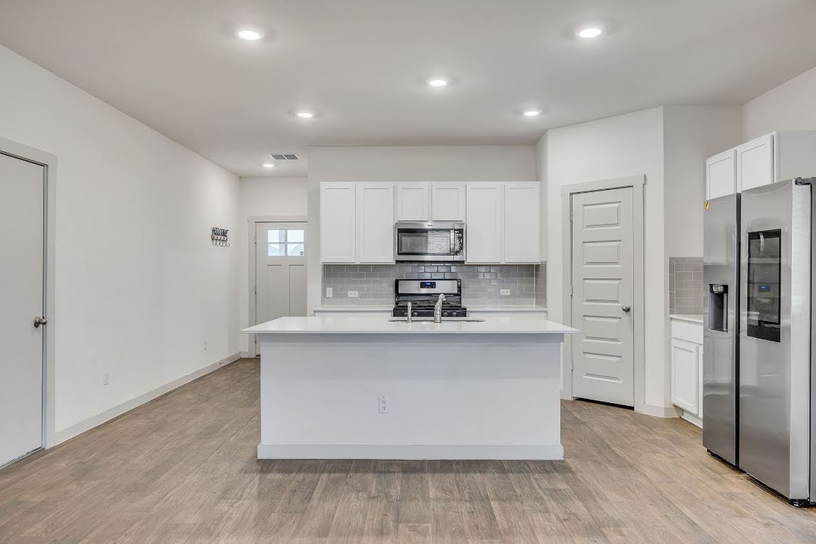 210 Big Cypress Bayou Road Hutto, TX 78634 - Photo 6 of 27 Kitchen featuring stainless steel appliances, white cabinetry, a kitchen island with sink, light wood finished floors, and backsplash