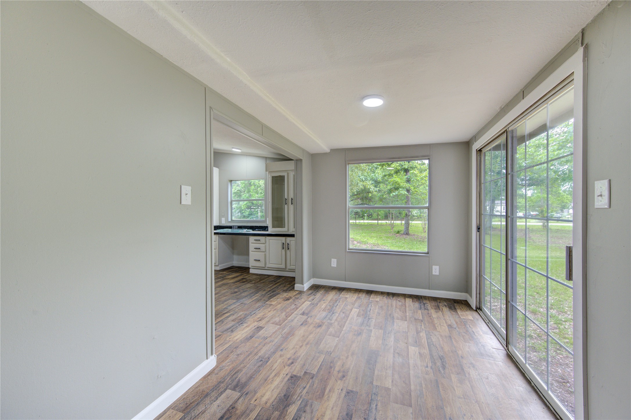 30226 Davis Street Magnolia, TX 77355 - Photo 13 of 50 This room features wooden flooring, neutral walls, and ample natural light from a large window and sliding glass doors leading to a view of greenery. Adjacent is a kitchen area with white cabinetry and a countertop.