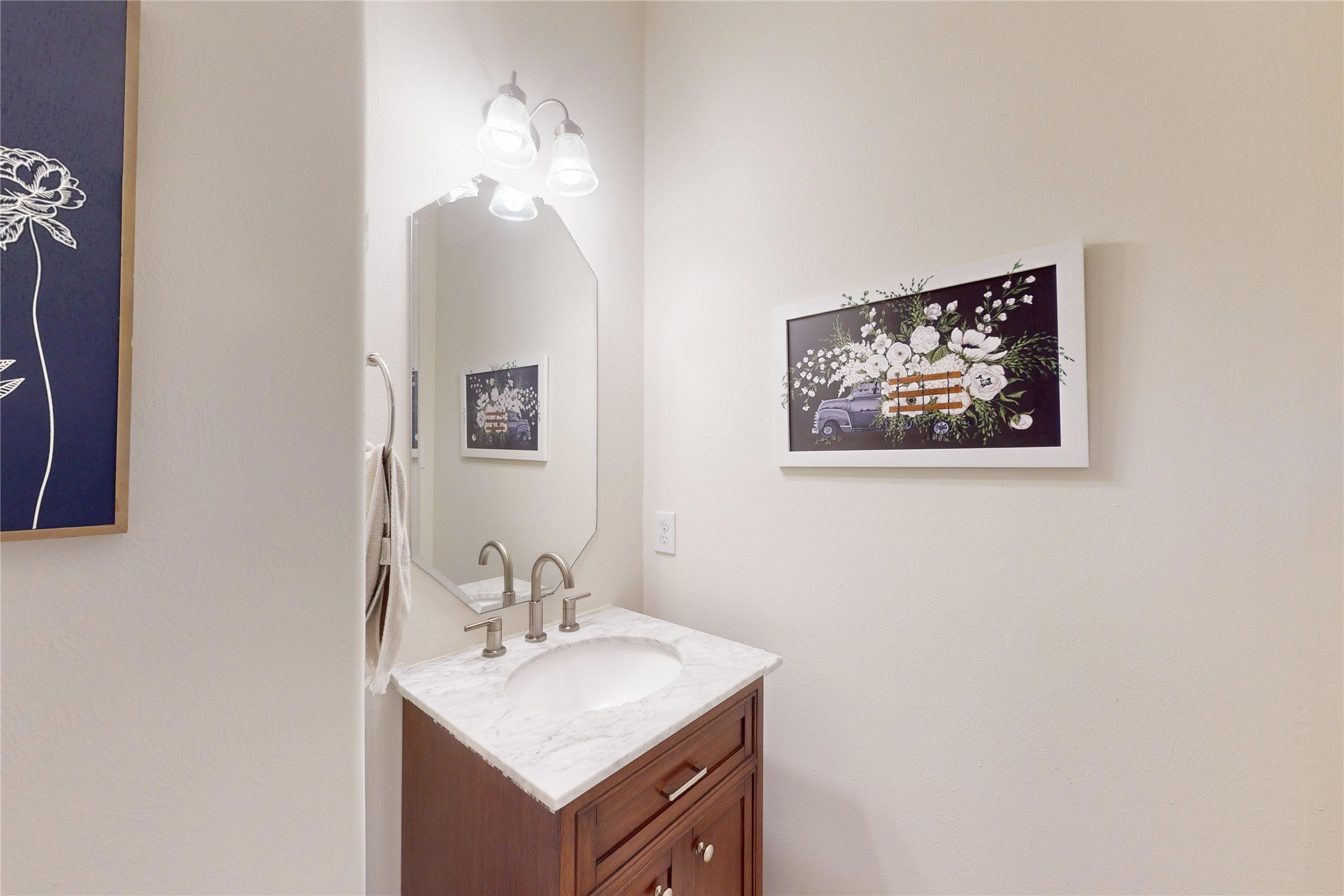 17402 Rainer Valley Lane Humble, TX 77346 - Photo 6 of 33 This photo shows a small, well-lit bathroom with a wooden vanity, marble style countertop, and modern fixtures. A mirror with overhead lighting is above the sink, and there's decorative art on the wall. The space is clean and contemporary.