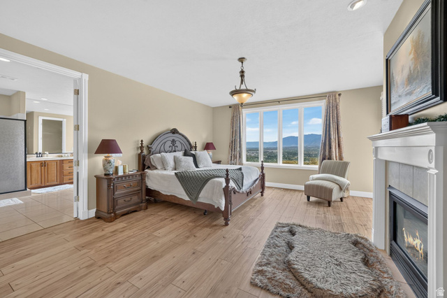 767 Draper View Road Draper, UT 84020 - Photo 23 of 71 Spacious bedroom featuring wood-finish flooring, a large window with mountain scenery, a white fireplace with a tiled surround, an ornate chandelier, and an adjoining bathroom with a glass-enclosed shower and wood vanity