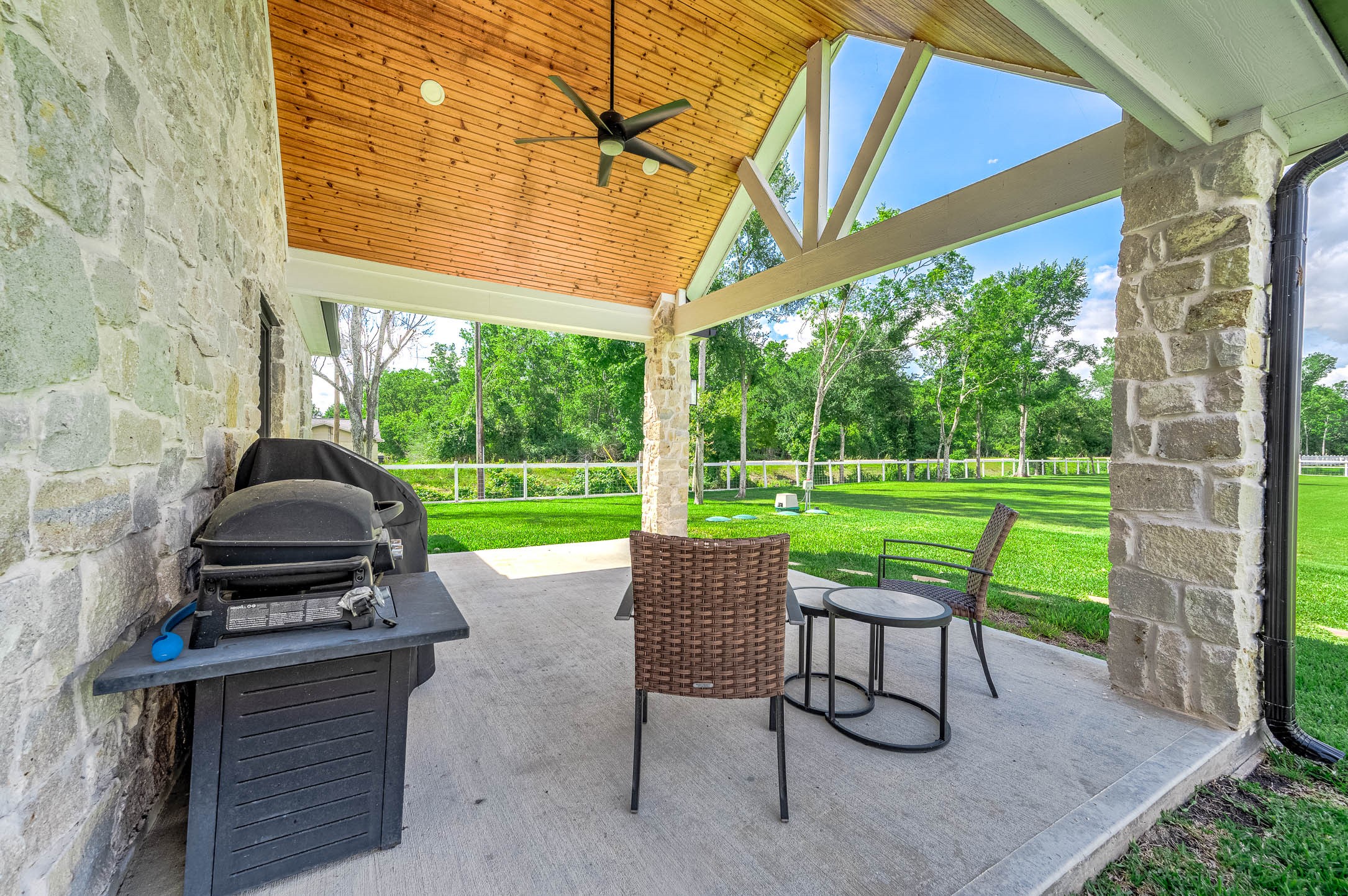 8032 Short Road Needville, TX 77461 - Photo 28 of 50 Enjoy outdoor living year-round on the covered back patio featuring a wood-accented ceiling, stone columns, and ceiling fan for added comfort. With plenty of room for seating and grilling, this space is perfect for relaxing or entertaining while overlooking the backyard.