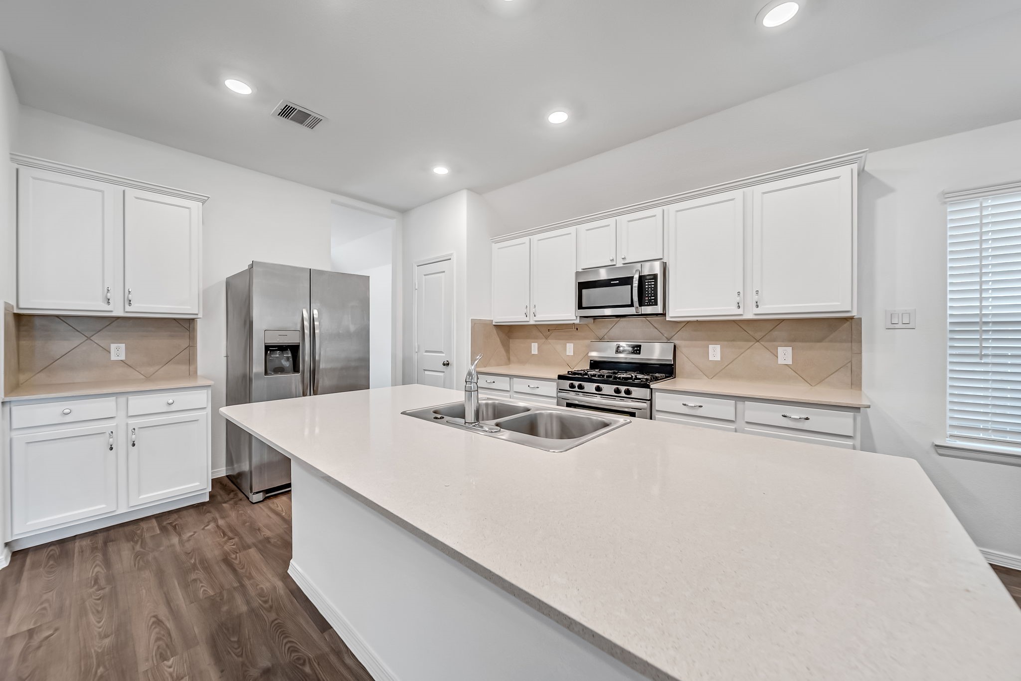 2847 Mistygate Court Conroe, TX 77301 - Photo 5 of 27 Modern kitchen with sleek white cabinetry, stainless steel appliances, and a spacious island featuring a double sink. The room is bright with recessed lighting and wood flooring, offering a clean and functional space.
