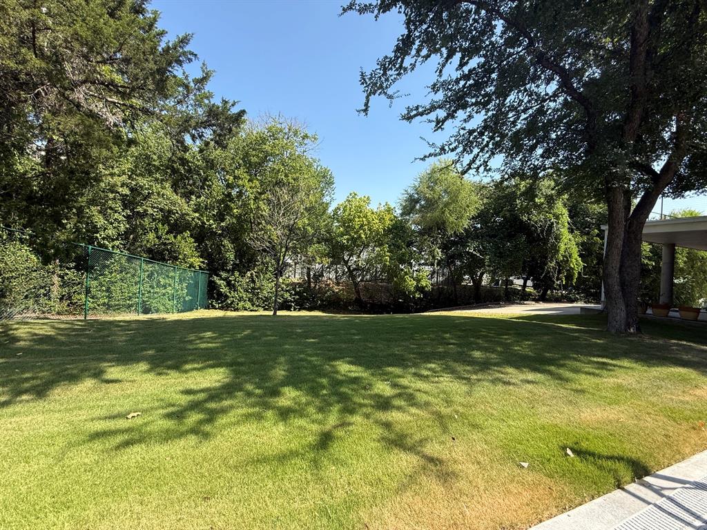 13023 Copenhill Road Dallas, TX 75240 - Photo 33 of 35 Expansive green lawn surrounded by mature trees, featuring a chain-link fence and a paved border