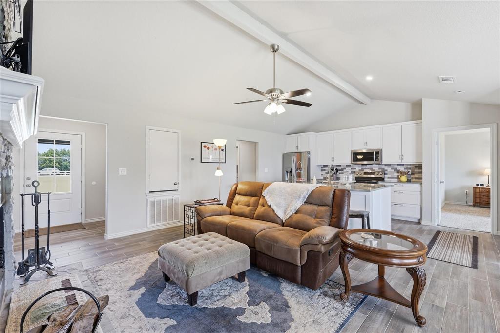 110 Highview Road Ennis, TX 75119 - Photo 8 of 40 Spacious living area featuring vaulted ceilings with exposed beams, wood-finish flooring, a stone-faced fireplace, and an open-concept kitchen with white cabinetry, stainless steel appliances, and a tiled backsplash