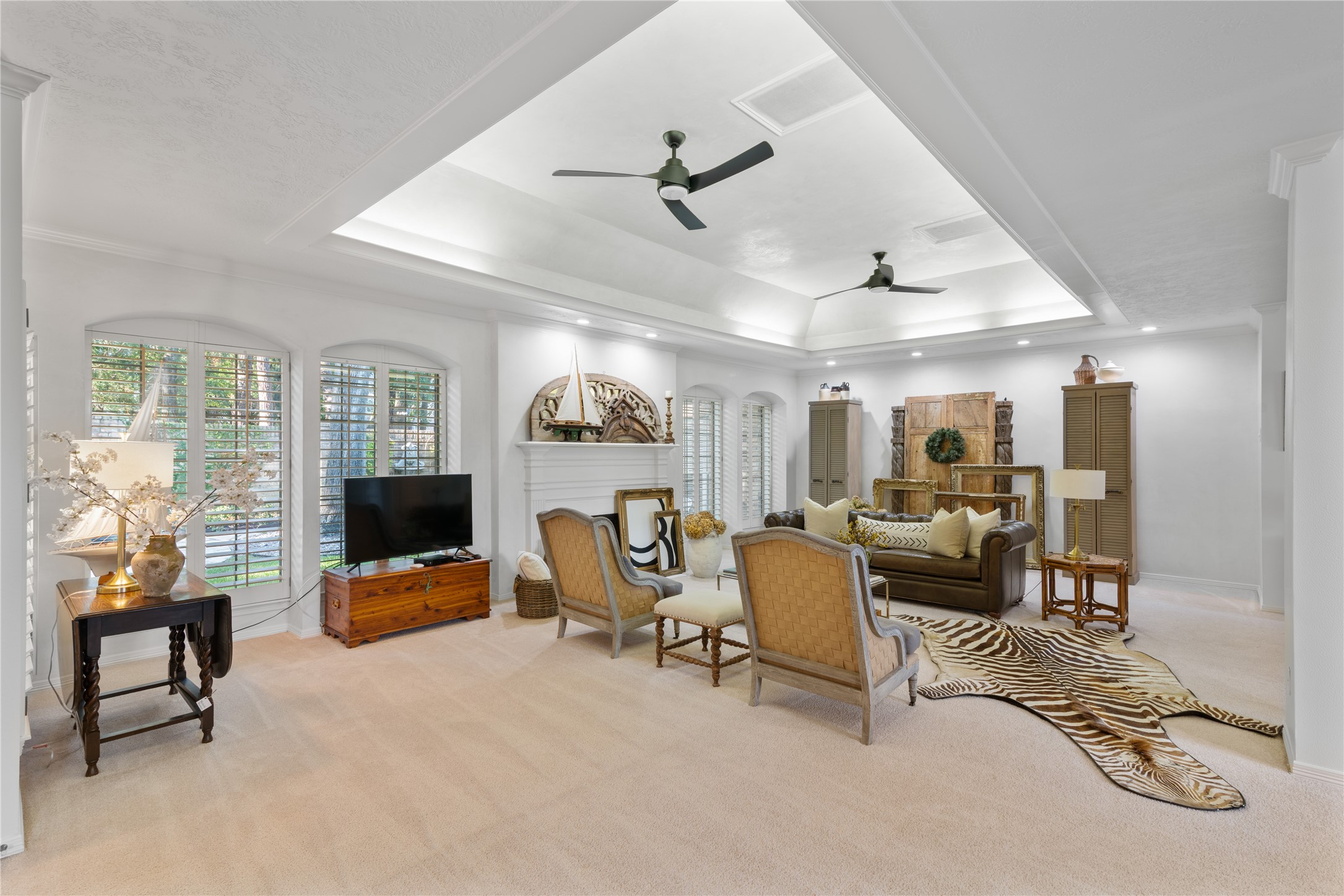 901 Longmire Road, Unit 51 Conroe, TX 77304 - Photo 4 of 48 Elegant living room featuring a coffered ceiling with recessed lighting and ceiling fans, complemented by a cozy seating area, a decorative fireplace, and large windows with plantation shutters, all creating a bright and inviting atmosphere.