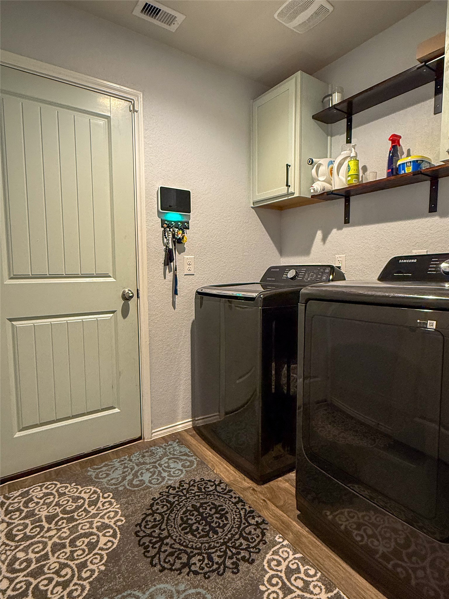 706 Zinnia Court Rosharon, TX 77583 - Photo 6 of 13 This photo shows a compact laundry room featuring a washer and dryer set against a wall, which are included in the sell of the home. There's shelving above for storage, a small cabinet, and a decorative rug on the floor. The room also has a key holder and a door leading out to the two car garage.