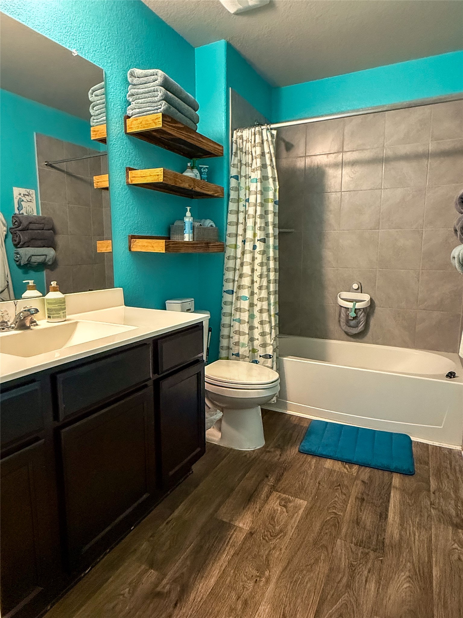 706 Zinnia Court Rosharon, TX 77583 - Photo 7 of 13 This bathroom features vibrant turquoise walls, a modern vanity with a large mirror, and wood-look flooring. It includes a bathtub with a shower and tile surround, and wooden shelves for storage.