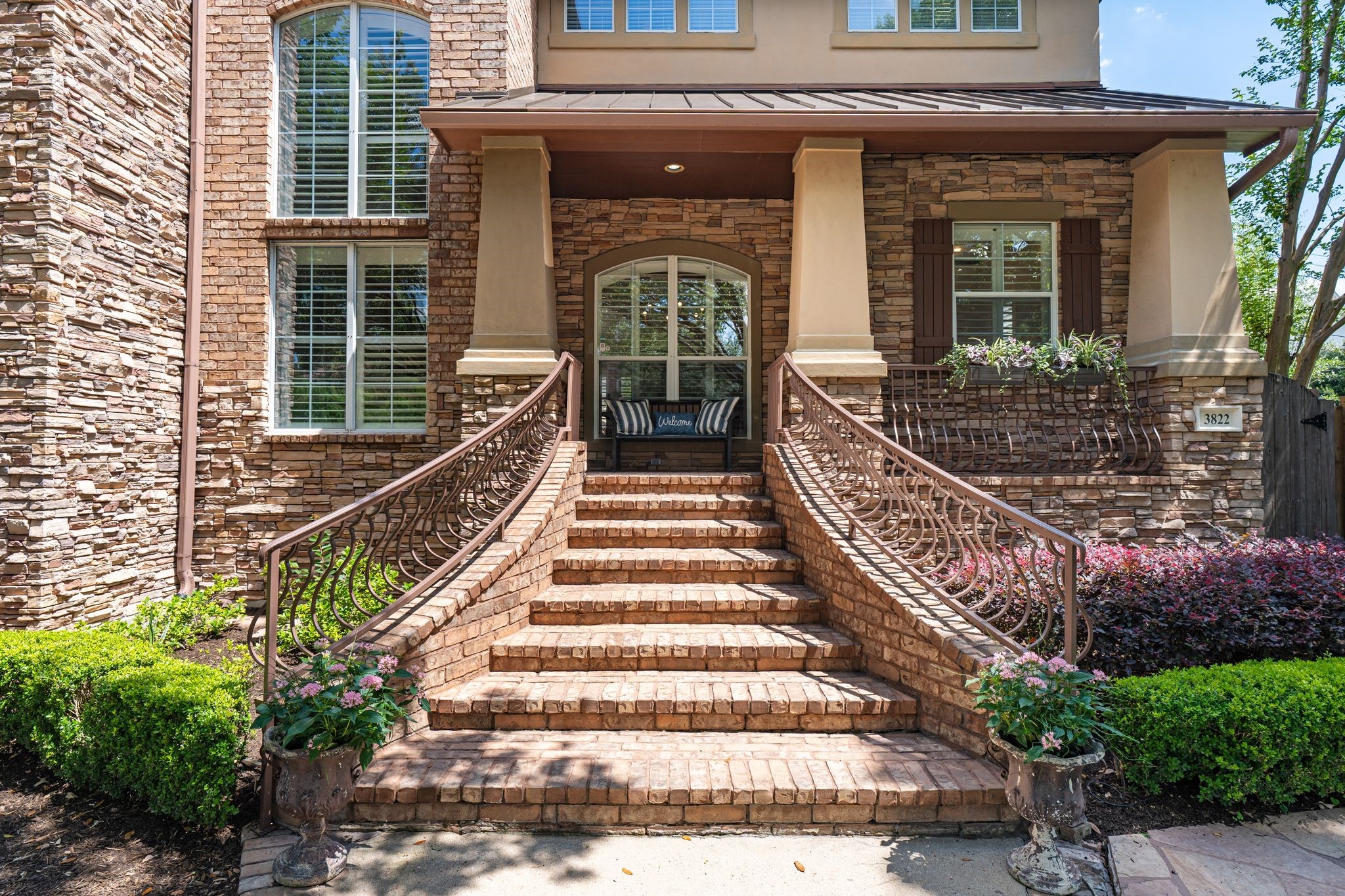 3822 Merrick Street Houston, TX 77025 - Photo 28 of 34 A close-up of the front entry reveals the meticulous craftsmanship of the brick stair treads, ornate curved iron railings with a custom bronze finish, stacked stone columns, and the arched covered porch entry above. The house number 3822 is displayed prominently on the stone façade, flanked by manicured boxwood hedges and colorful plantings.