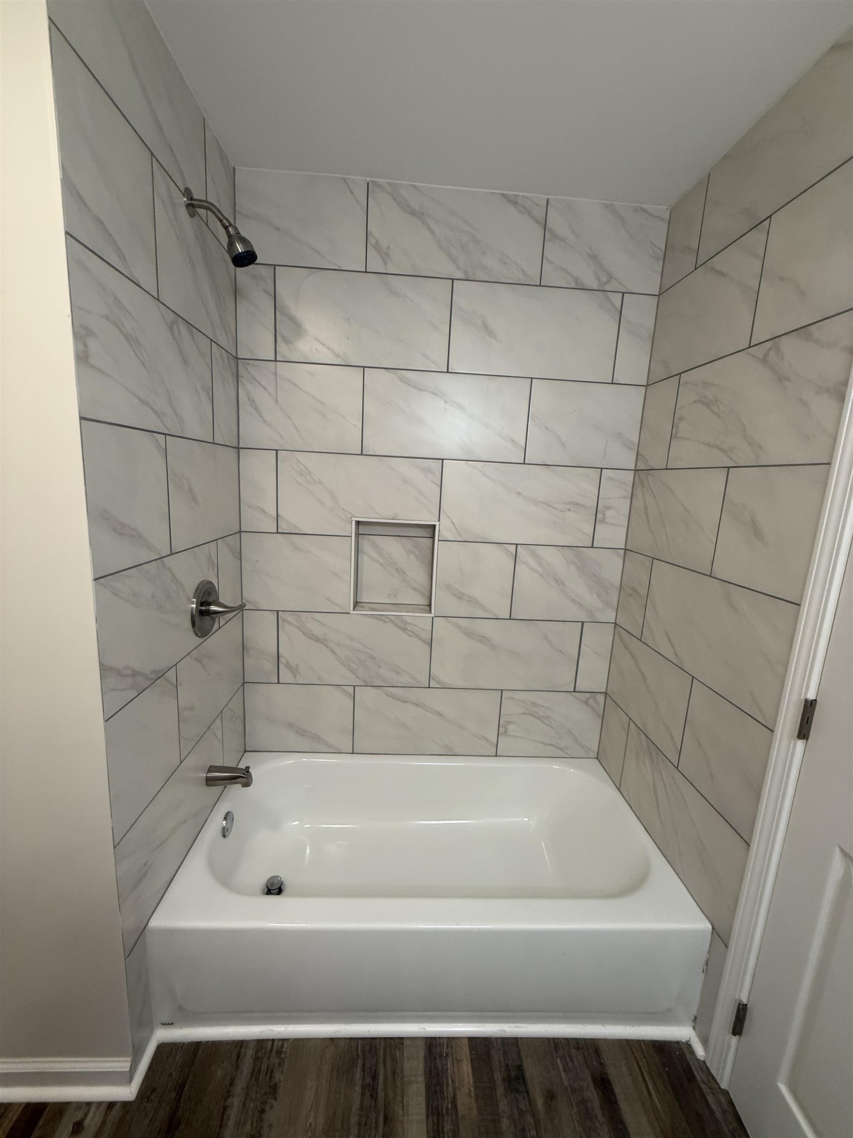 1835 Pleasant Grove Road Moscow, TN 38057 - Photo 23 of 34 Bathtub with integrated shower featuring white subway tiles with subtle gray veining, chrome fixtures, a recessed shower niche, and wood-finish flooring