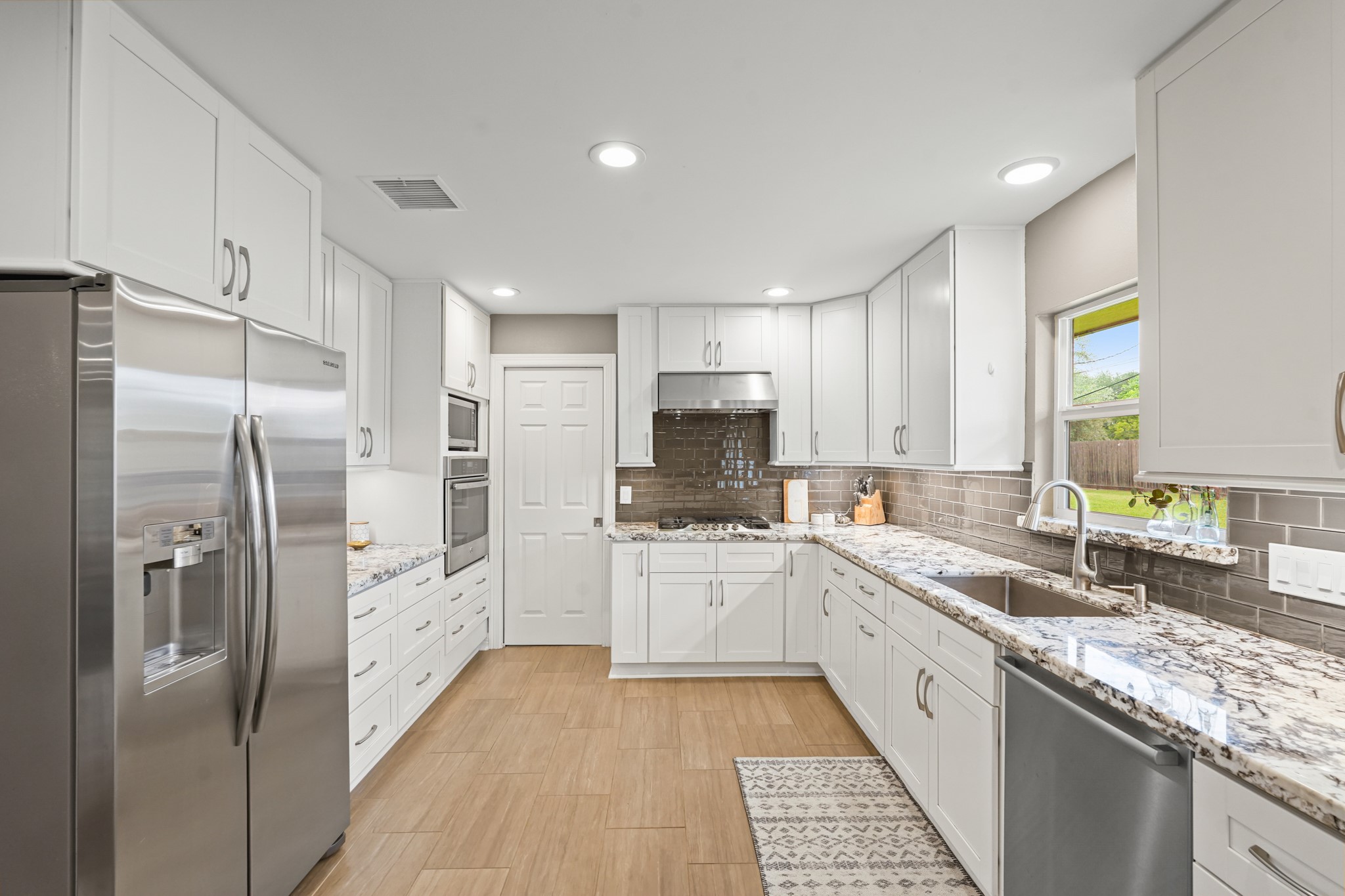 5242 Sanford Road Houston, TX 77035 - Photo 12 of 34 This modern kitchen features sleek stainless steel appliances, abundant white cabinetry, and elegant granite countertops. It has a spacious layout with a double sink, a built-in oven, and a large fridge, all complemented by stylish tile flooring and a subway tile backsplash. Perfect for home cooking and entertaining.