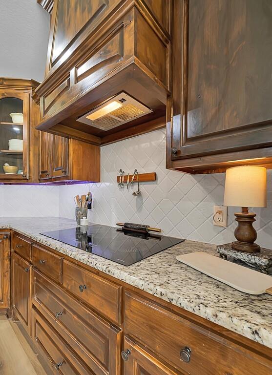 550 Hunton Lane Robinson, TX 76706 - Photo 12 of 35 Kitchen with custom range hood, backsplash, light stone counters, black electric cooktop, and glass insert cabinets
