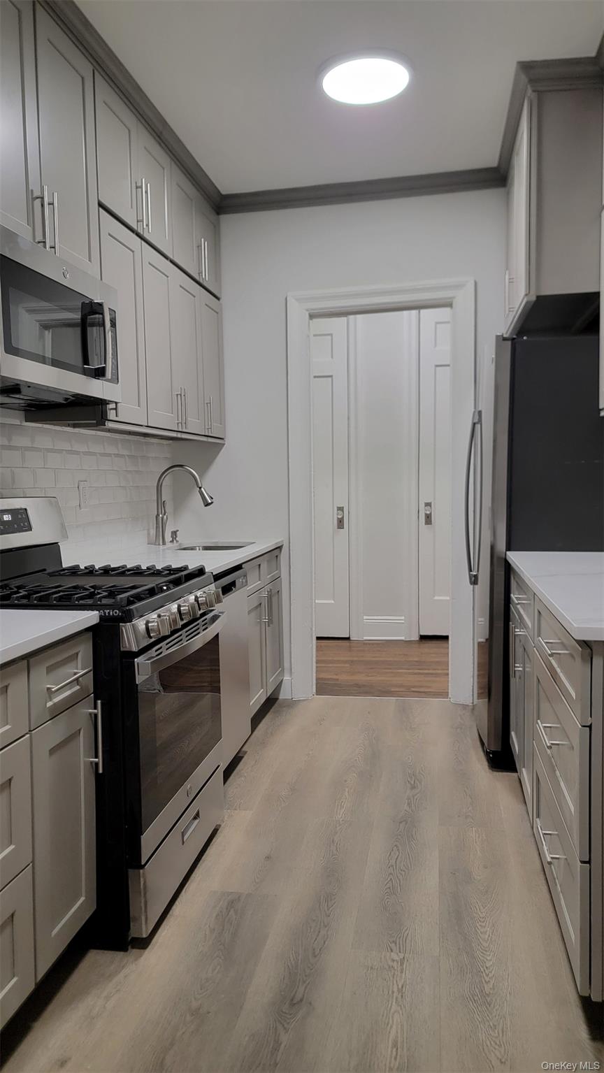 34-58 90th Street, Unit R5 Queens, NY 11372 - Photo 9 of 14 Kitchen with gray cabinets, appliances with stainless steel finishes, light wood finished floors, backsplash, and light stone counters