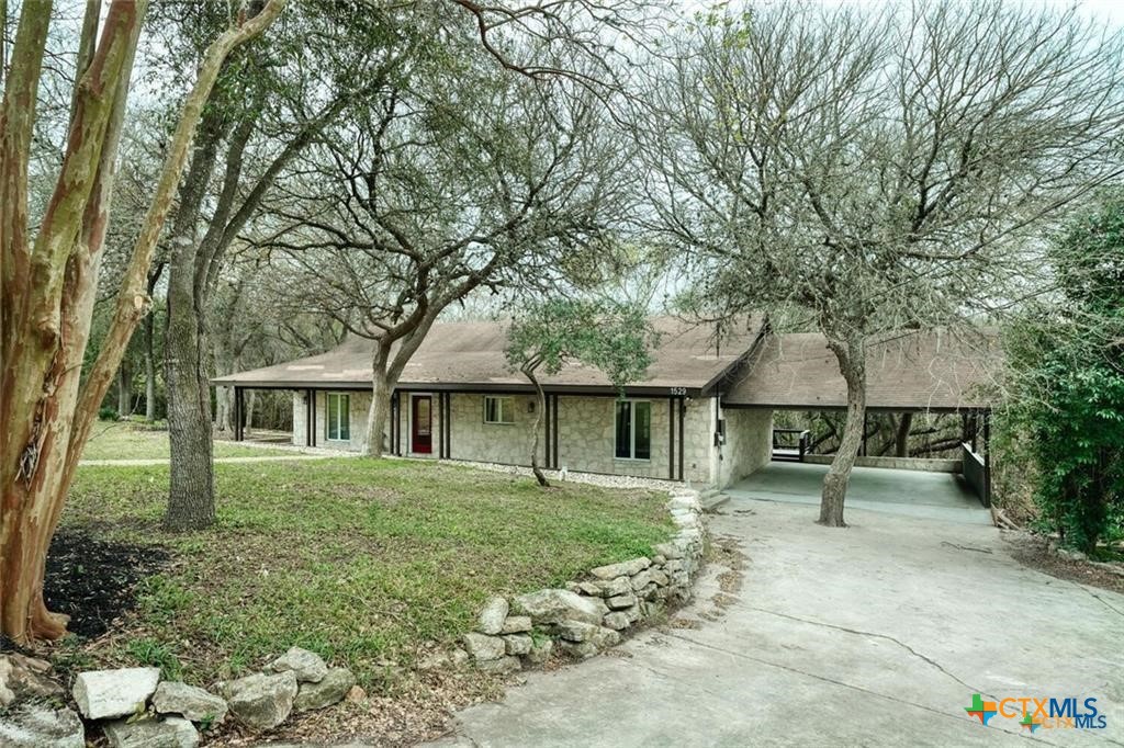 1529 North Colorado Street Lockhart, TX 78644 - Photo 1 of 14 View of the front of the home shows the mature trees, large lot with long driveway leading to a large 2-car carport. Stone covers the home on three sides.