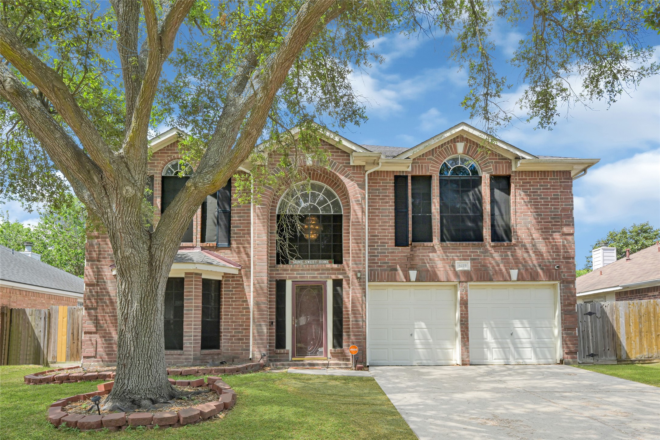 This two-story brick home features a spacious front yard with a mature tree, a two-car garage, and a prominent arched window above the entrance, offering great curb appeal.
