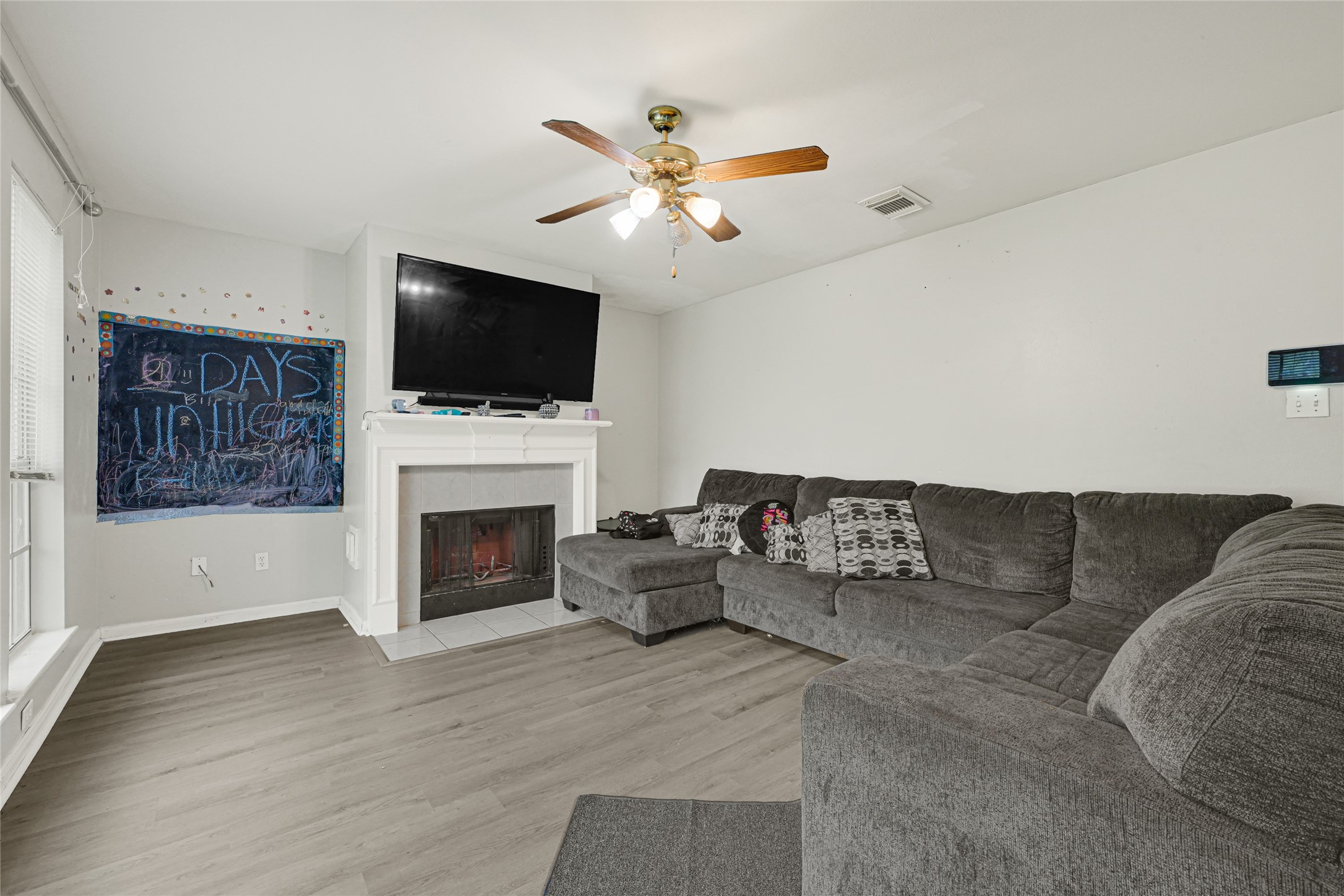 6419 Slate Stone Street Houston, TX 77084 - Photo 15 of 49 Cozy living room with a large sectional sofa, fireplace, and mounted TV. Features a ceiling fan, chalkboard wall, and wood-style flooring, offering a welcoming and functional space for relaxation.