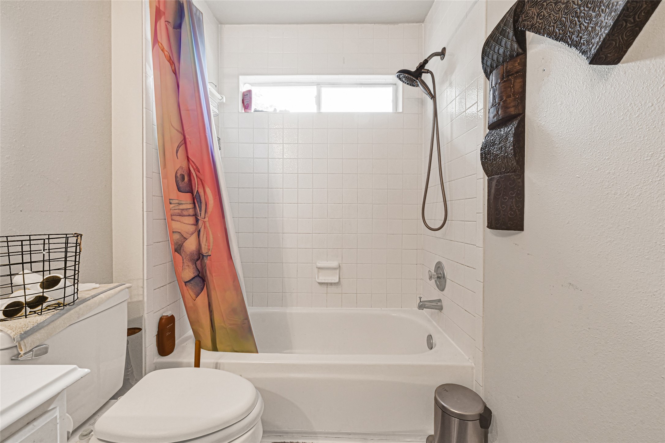 6419 Slate Stone Street Houston, TX 77084 - Photo 25 of 49 This bathroom features a white-tiled bathtub and shower combo with a window for natural light. It includes a colorful shower curtain, modern fixtures, and a nearby toilet. The decor is minimal with a small basket on the countertop and wall art for added style.
