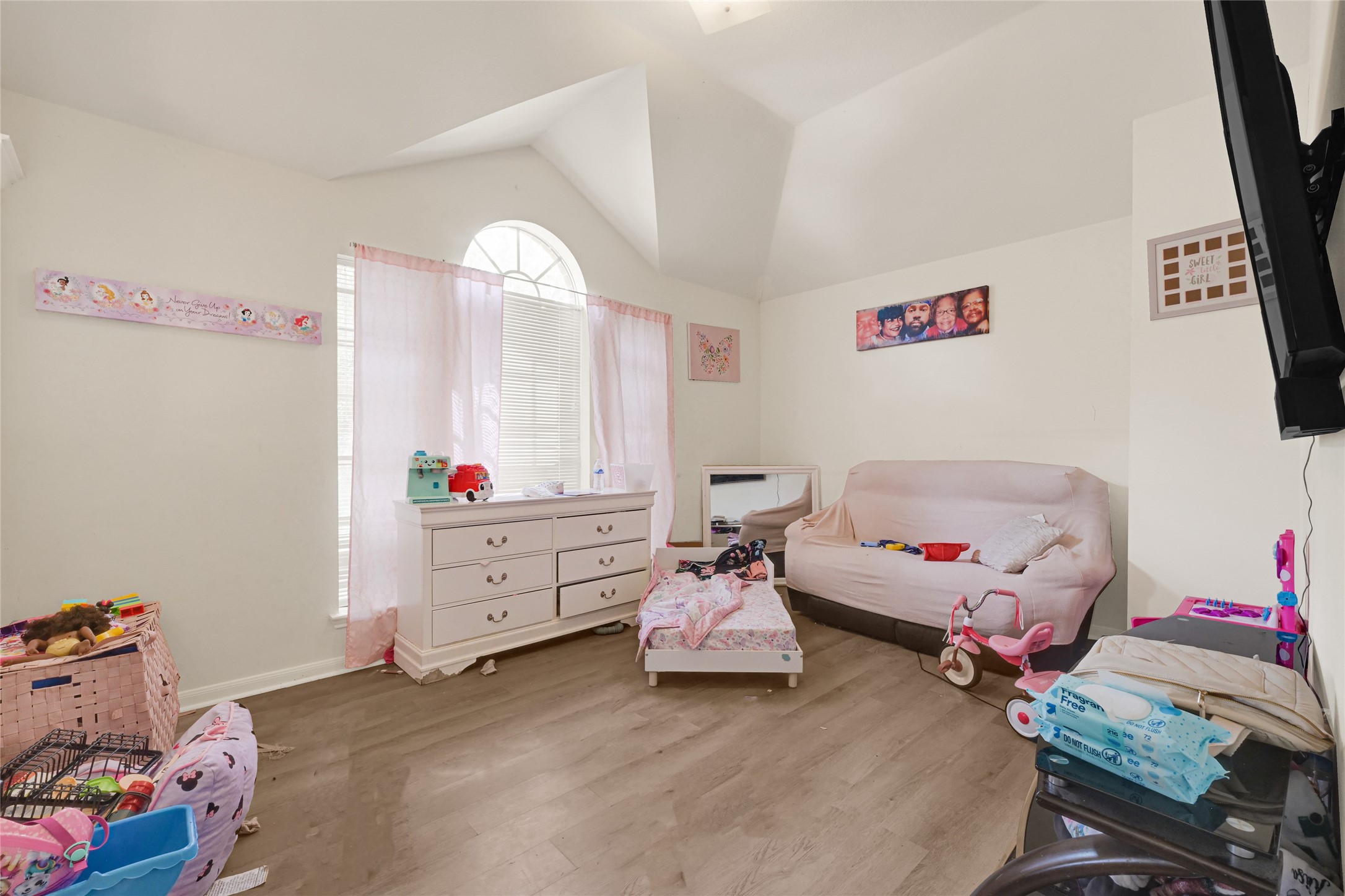 6419 Slate Stone Street Houston, TX 77084 - Photo 33 of 49 This cozy room features a vaulted ceiling, large window with soft pink curtains, and hardwood floors. It includes a dresser, a small bed, and a sofa, making it ideal for a child's bedroom or playroom.